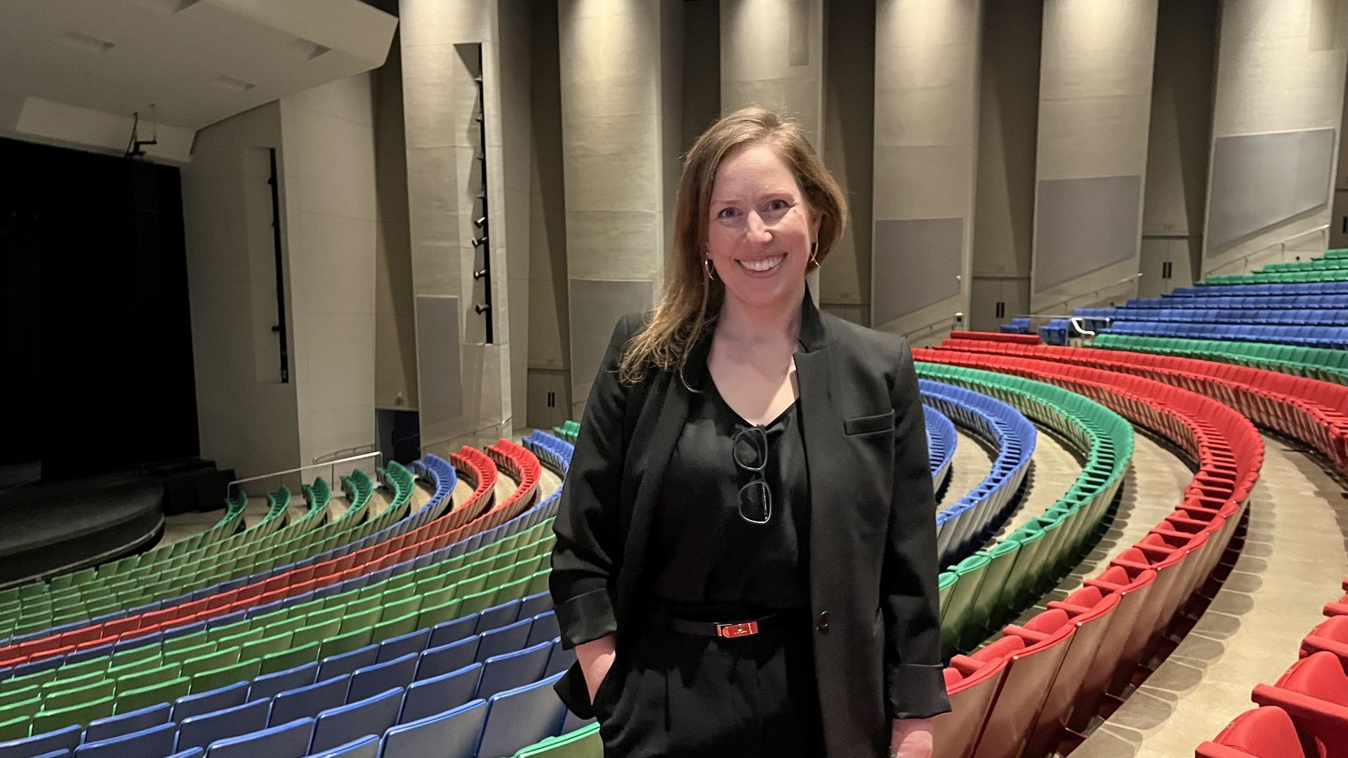 Smiling woman in a black blazer and top stands in a theater with curved, color-coded seating in green, blue, and red, and a stage on the left.