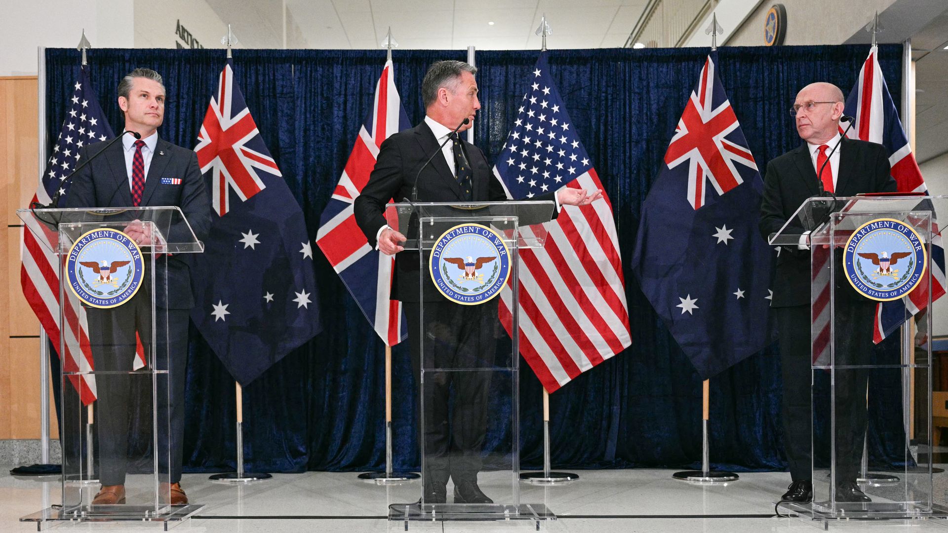 Three men in dark suits stand behind clear lecterns. The American, Australian and British flags hang behind them, against a blue curtain.