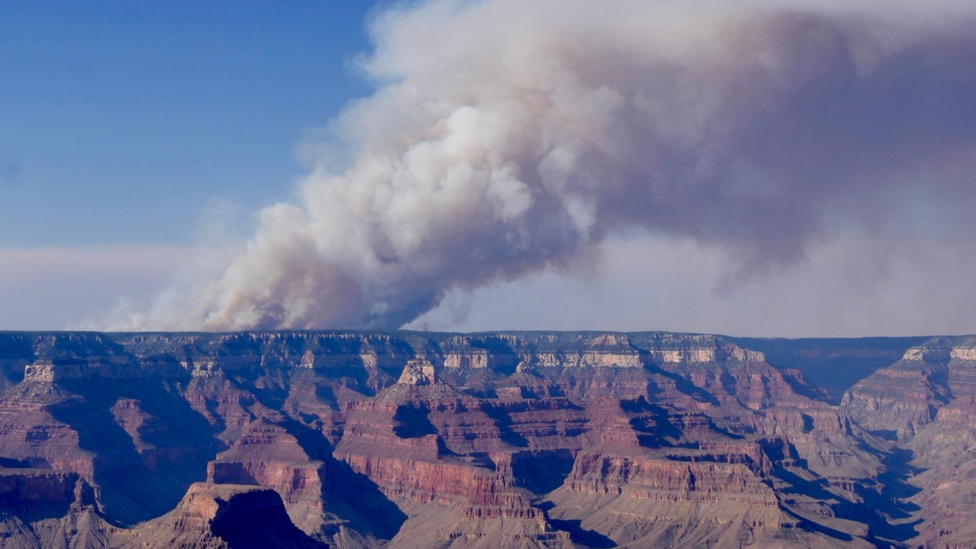 White and gray smoke billows up from the horizon, with the Grand Canyon in the foreground.