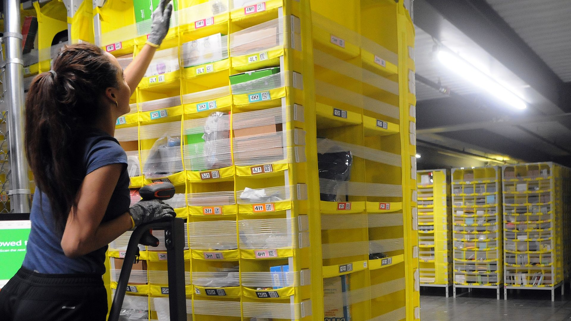 A woman reaches for an item on a yellow shelf in a warehouse
