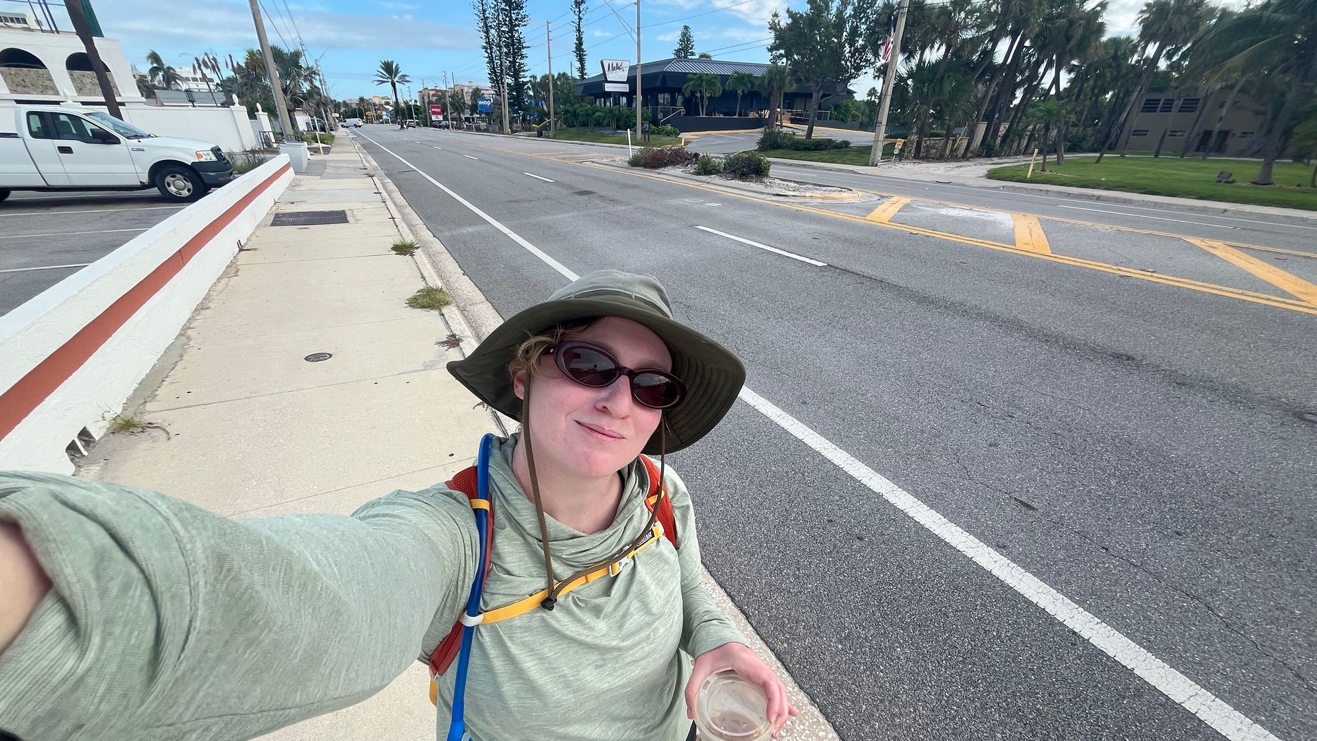 Person wearing a green long-sleeve shirt, wide-brimmed hat, and sunglasses takes a selfie on a sidewalk next to a mostly empty street, holding a plastic cup with iced coffee.