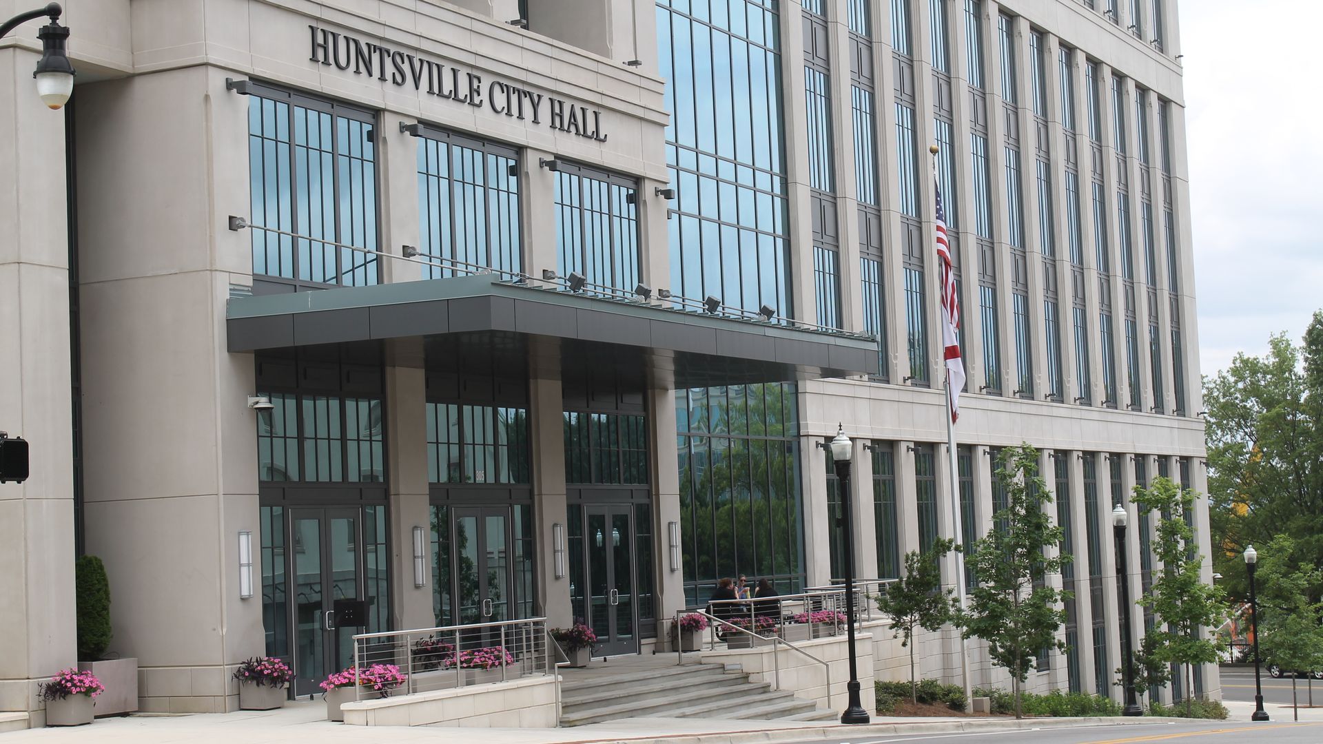 Modern Huntsville City Hall building with large glass windows, beige stone exterior, entrance canopy, flower pots, American flag, and street lamps along the sidewalk.