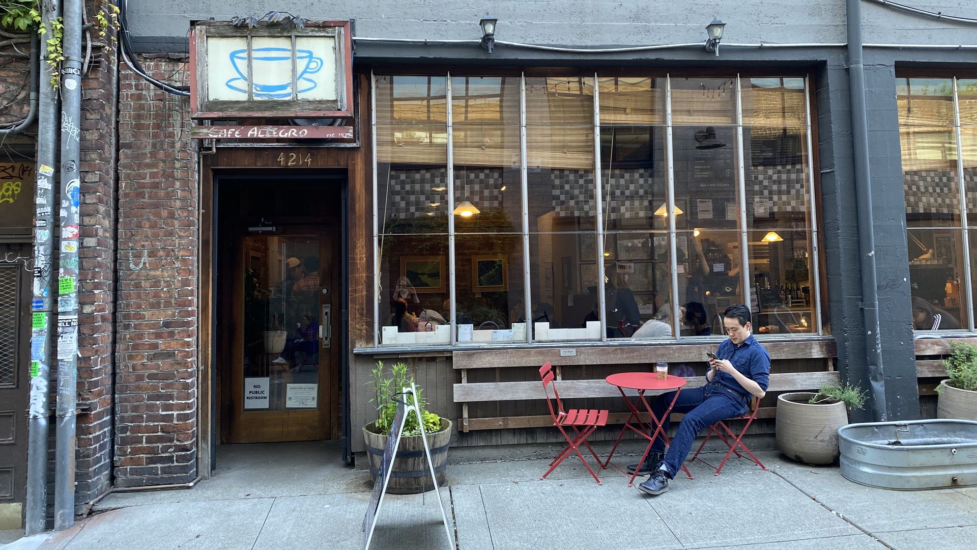 A brick facade with large windows and a hanging sign with a painted coffee mug that says "Cafe Allegro," with a sandwich board out front and a man sitting at a red cafe table. 
