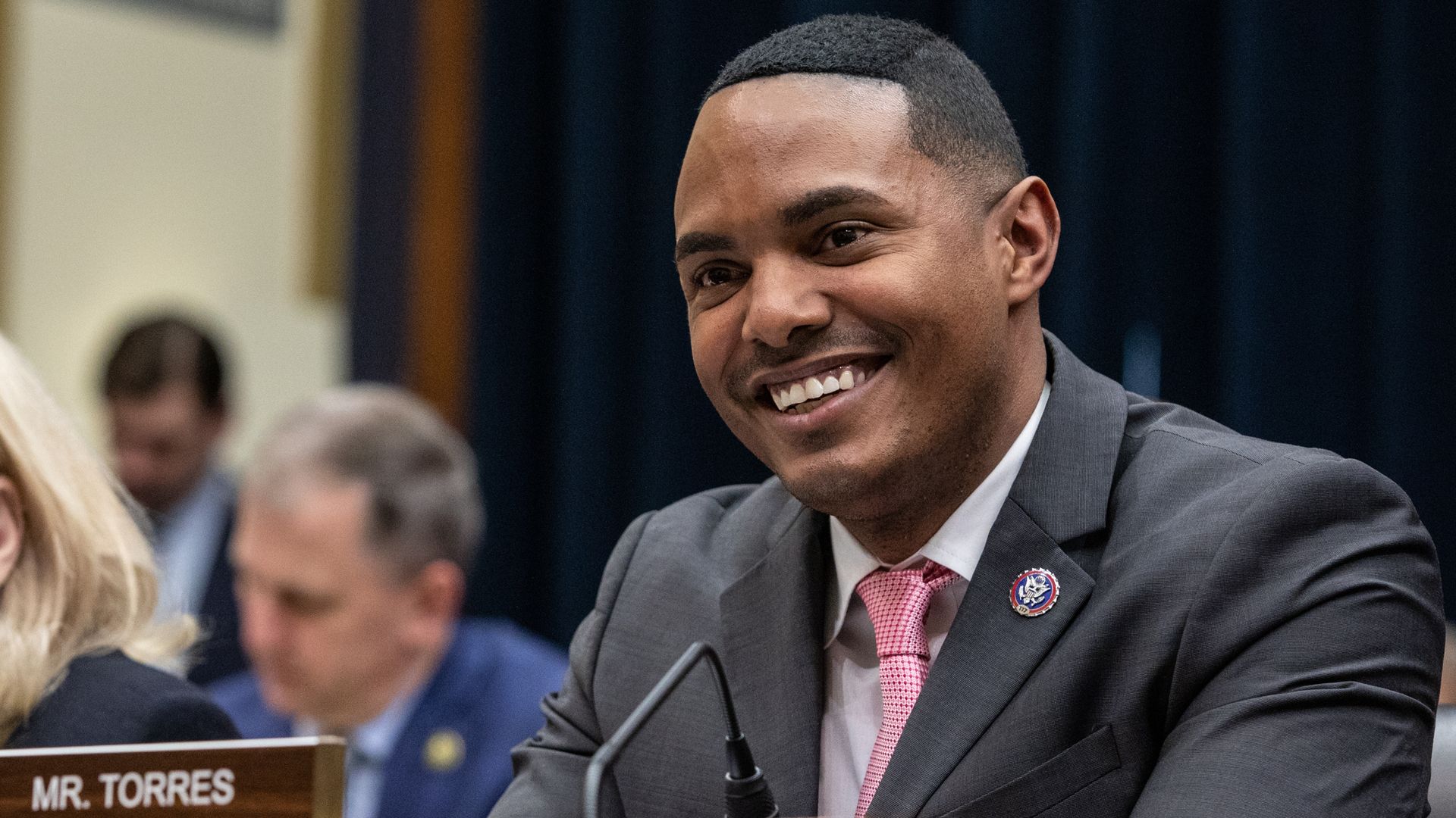 A man speaking at a microphone. Close-cropped hair, gray suit, pink tie. A few others blurry in the background.