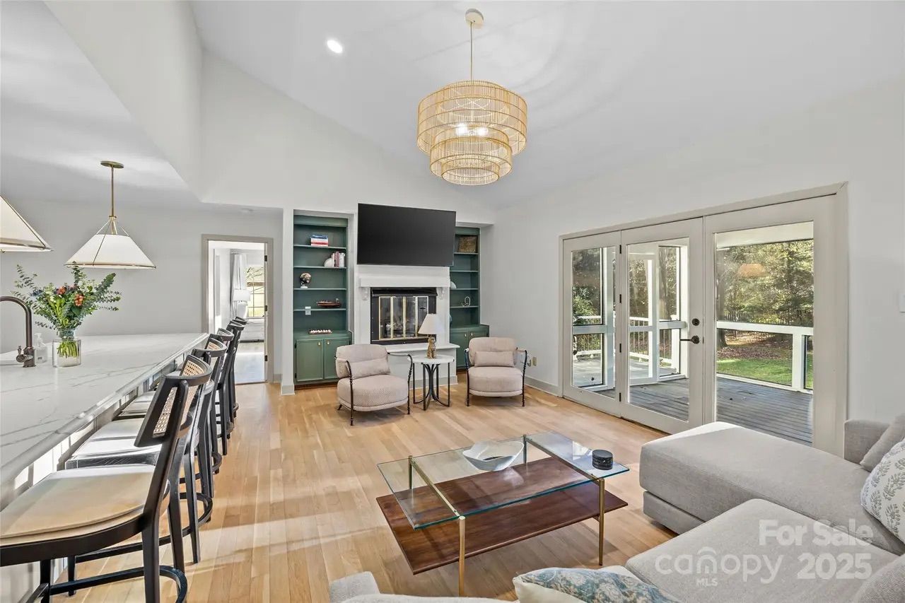 Bright living room with light wood floors, green built-in shelves, two armchairs by a fireplace, large glass doors opening to a porch, a modern pendant light, and a marble breakfast bar.