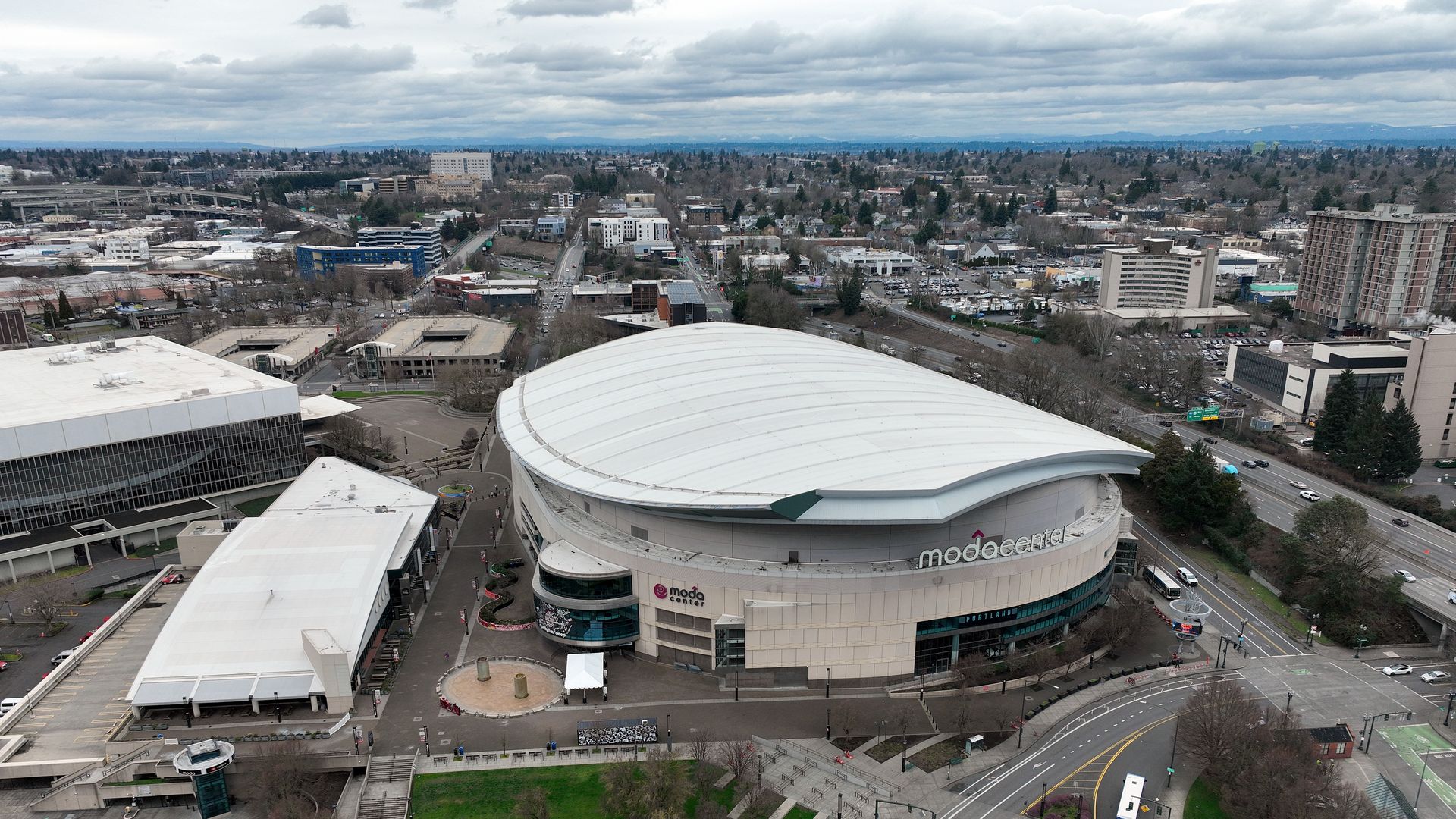 Aerial view of downtown with the Moda Center's large curved white roof in the foreground, surrounded by roads, midrise buildings, trees, and a cloudy sky.