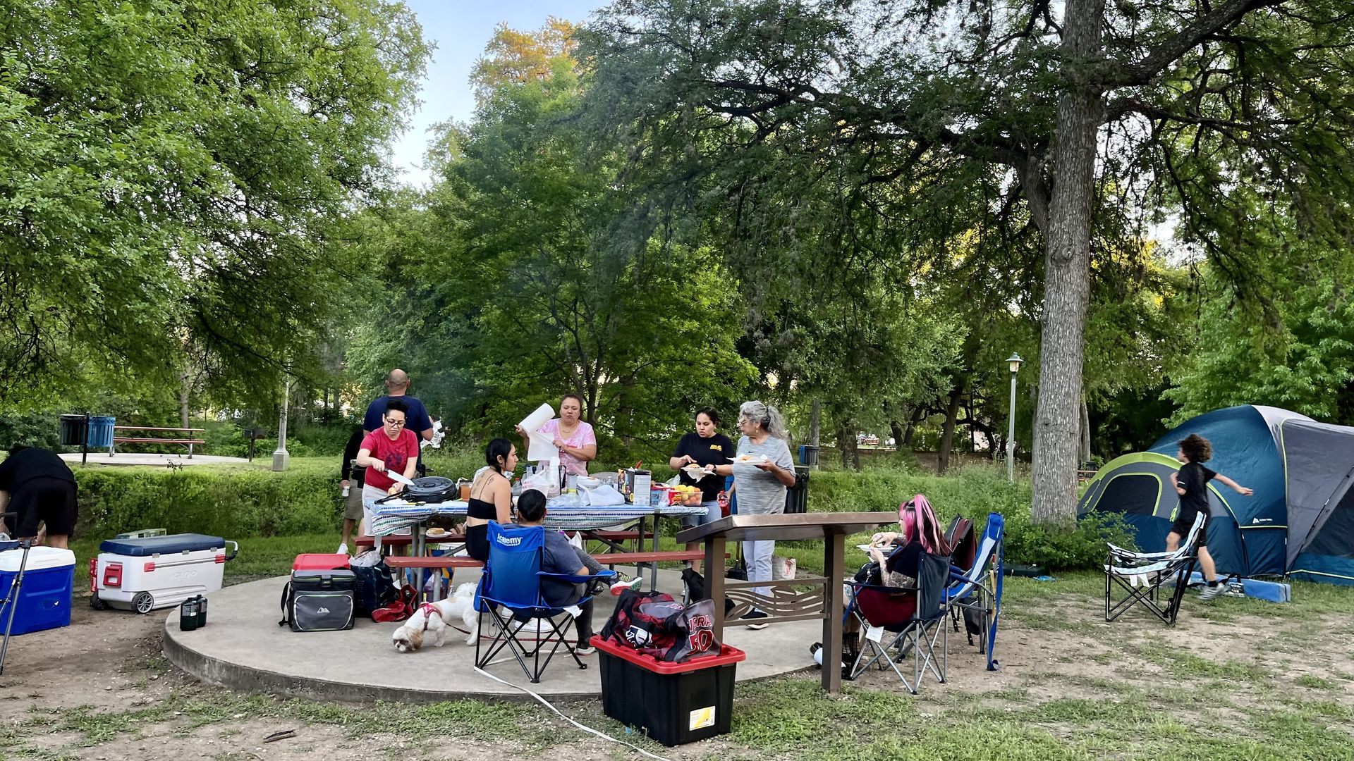 A family at a campsite around a picnic table, with a tent in the corner and a kid running past it. A dog is on a leash in the middle.