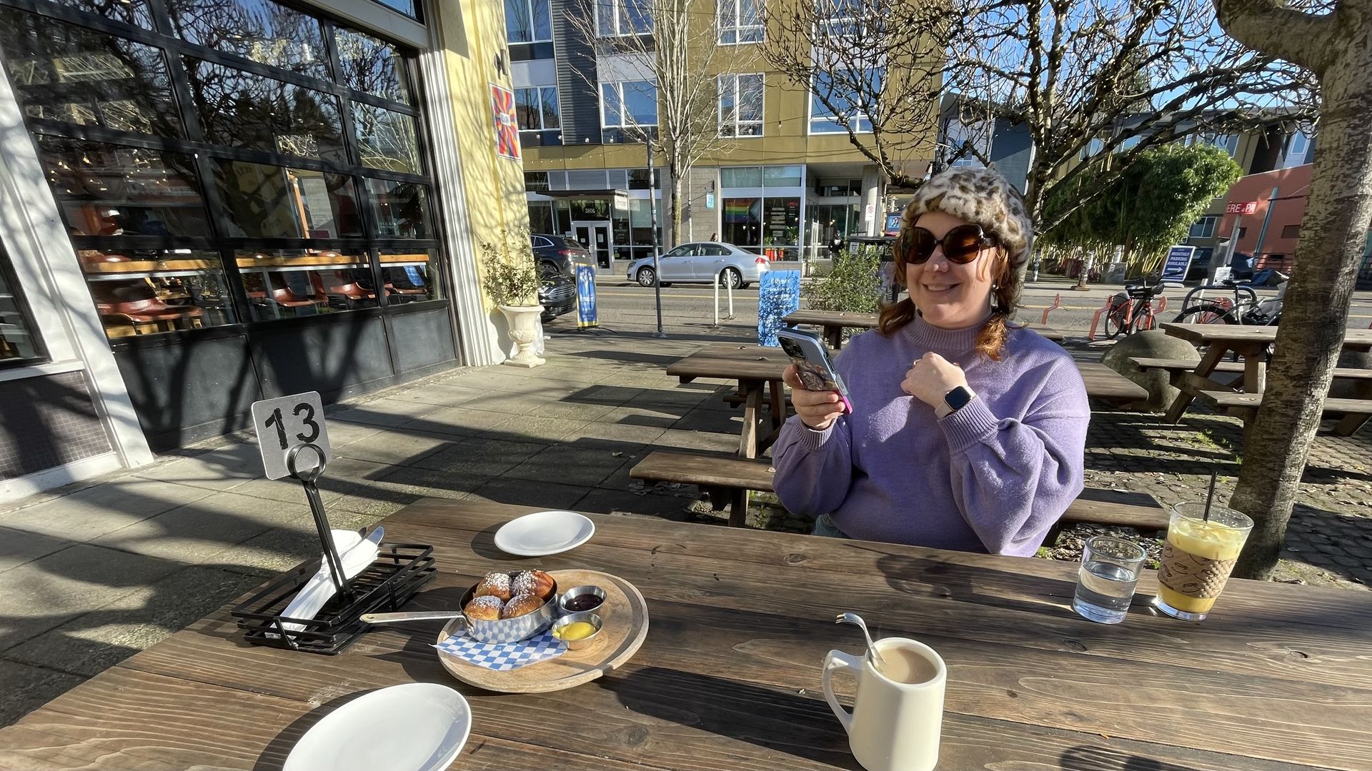 Woman in a purple sweater and leopard print hat sitting at an outdoor wooden table with pastries, coffee, and iced drink on a sunny day in an urban cafe setting.