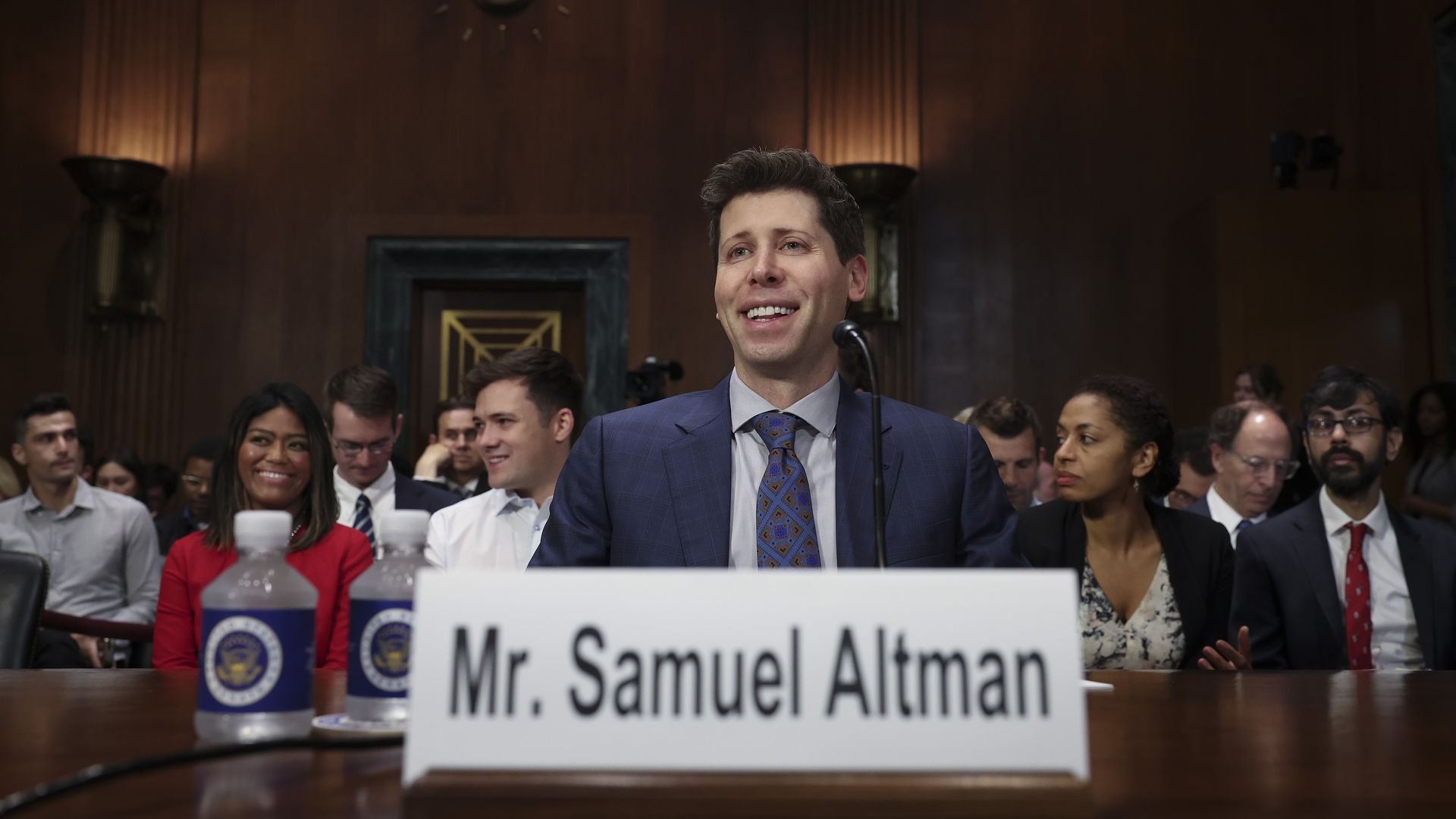 Photo of Sam Altman in suit and tie smiling, seated at testimony table, with a placard bearing his name