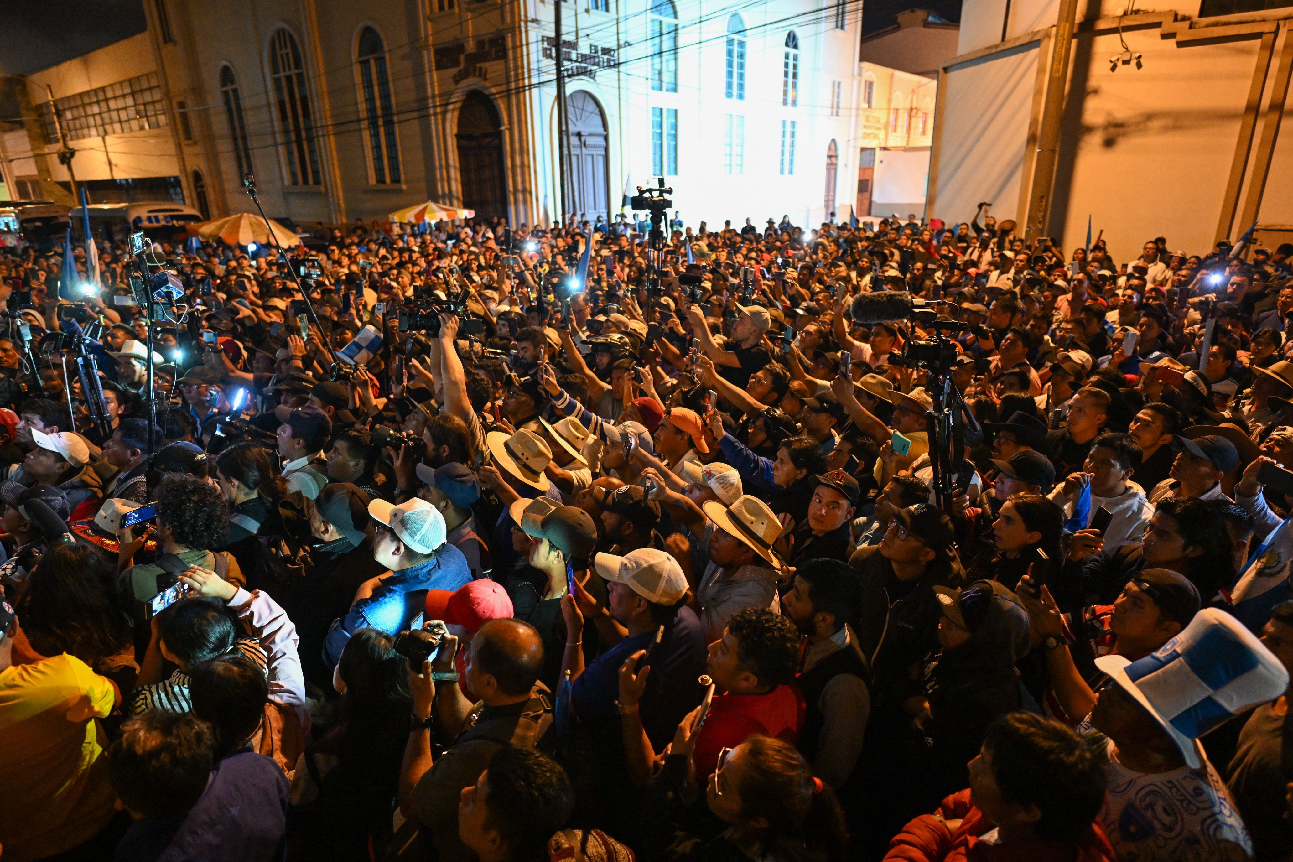 dozens of protestors stand outside Guatemalan government offices during the night, waving their arms in the air and holding up cell phone flashlights