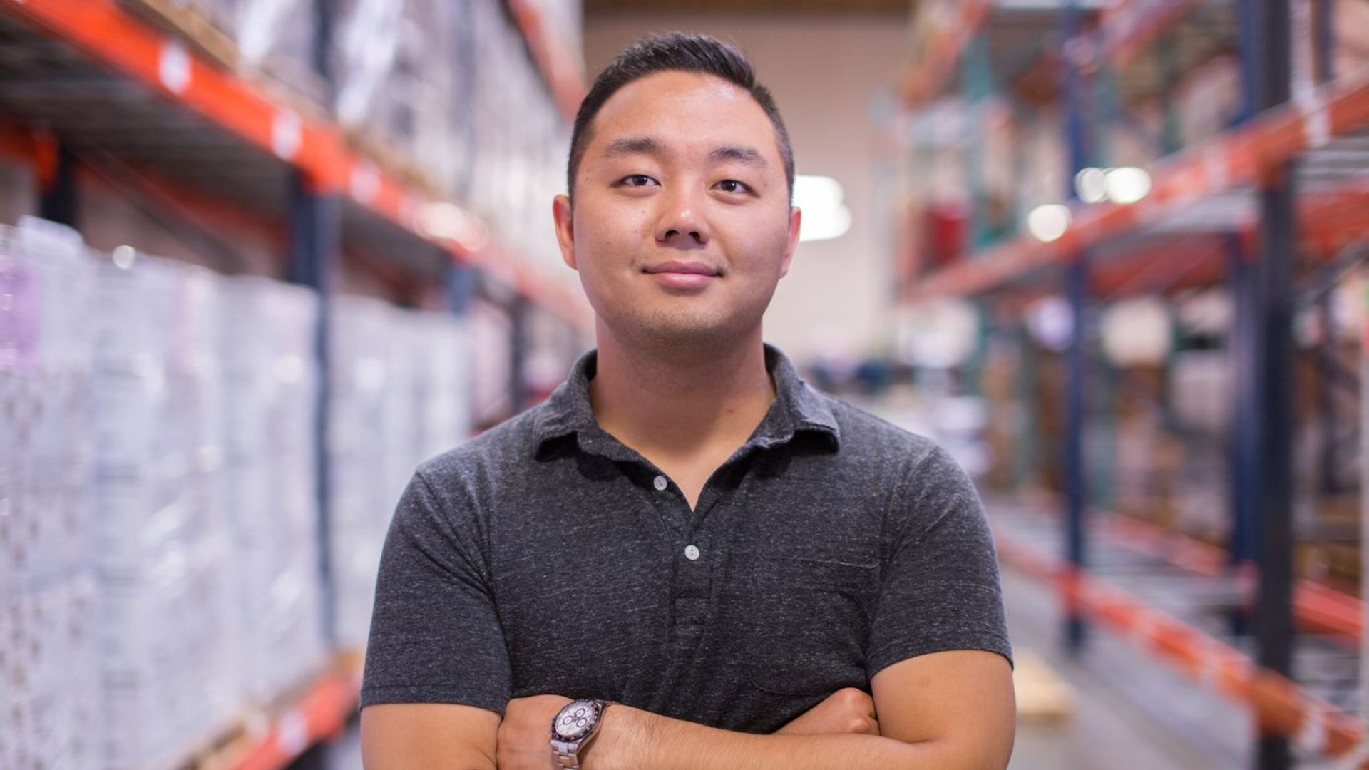 Eugene Kang, the founder of meat snacks brand Country Archer, stands in the middle of his distribution facility with arms crossed wearing a grey polo shirt.