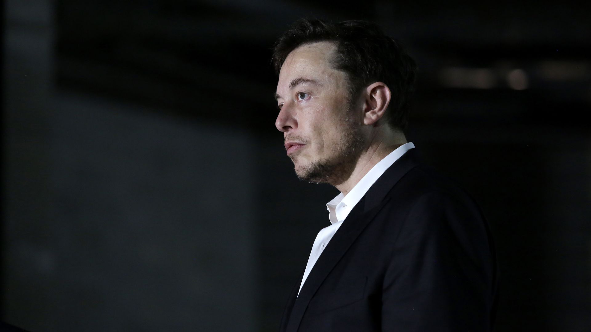 Elon Musk from SpaceX in a dark room. He is standing with his profile to the camera and pursing his lips. 