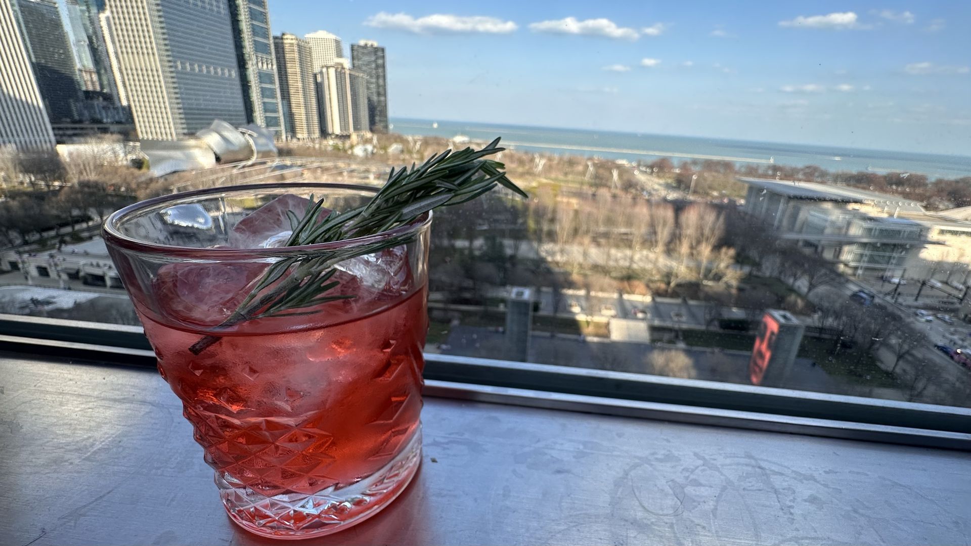 Red cocktail in a faceted glass with ice and a rosemary sprig, set on a windowsill. Behind it is the Chicago skyline and a calm blue bay under a clear sky.
