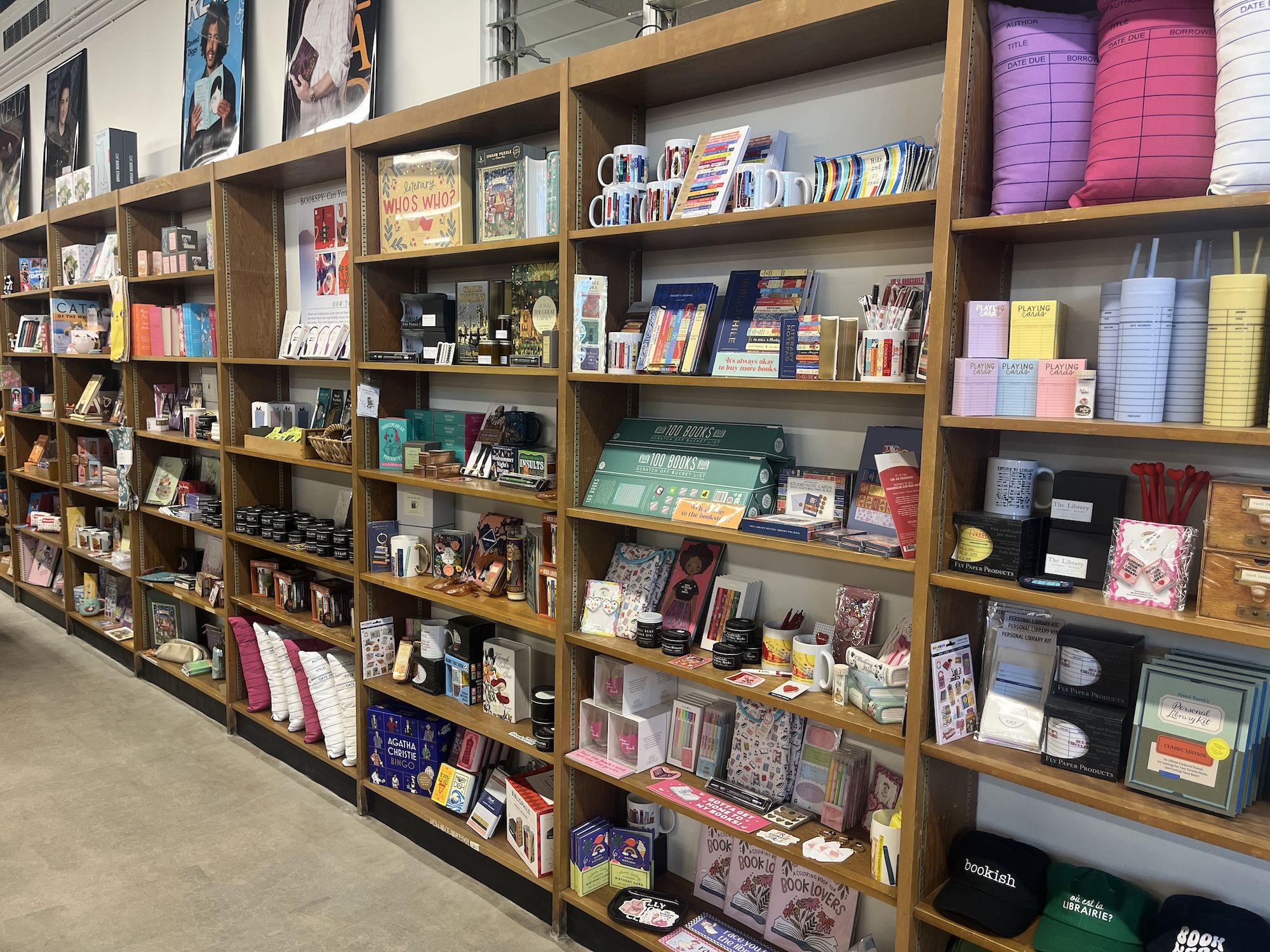 Wooden shelves filled with colorful books, mugs, pillows shaped like library due date cards, stationery, and various bookish merchandise in a store.