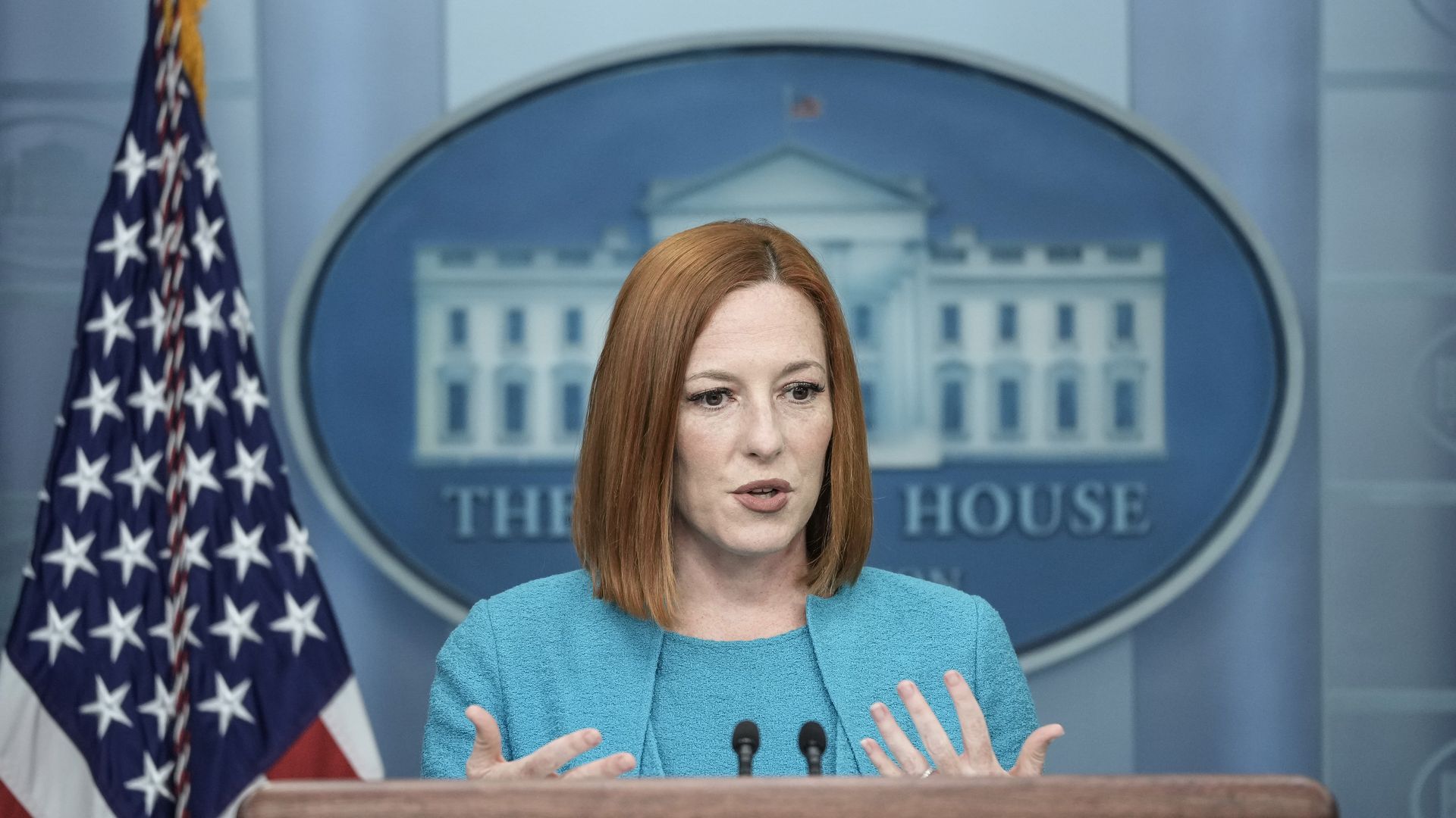 Jen Psaki speaks at a microphone with a U.S. flag and the White House logo in the background.