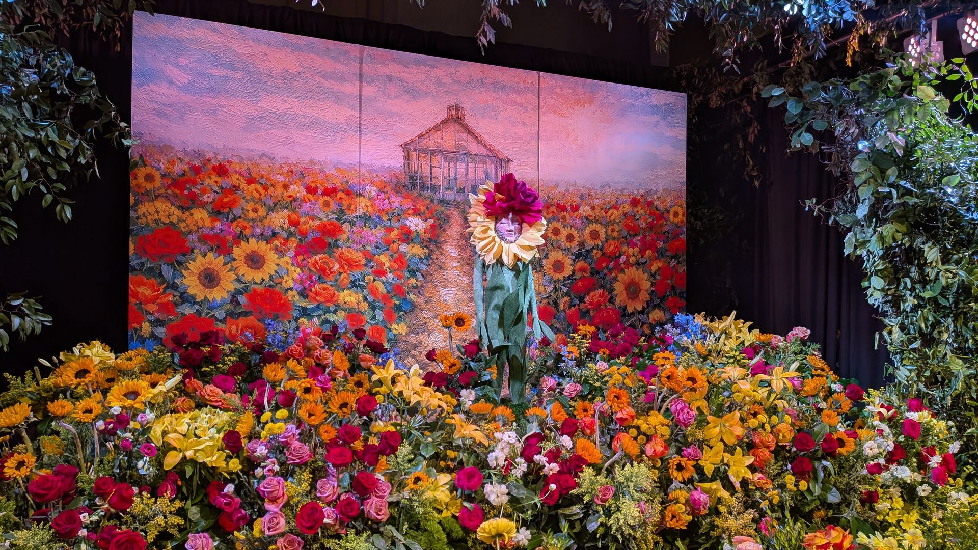 Three-panel mural of a sunlit field of flowers with a small greenhouse in the distance; foreground blooms surround a figure with a sunflower headdress, framed by leafy plants.