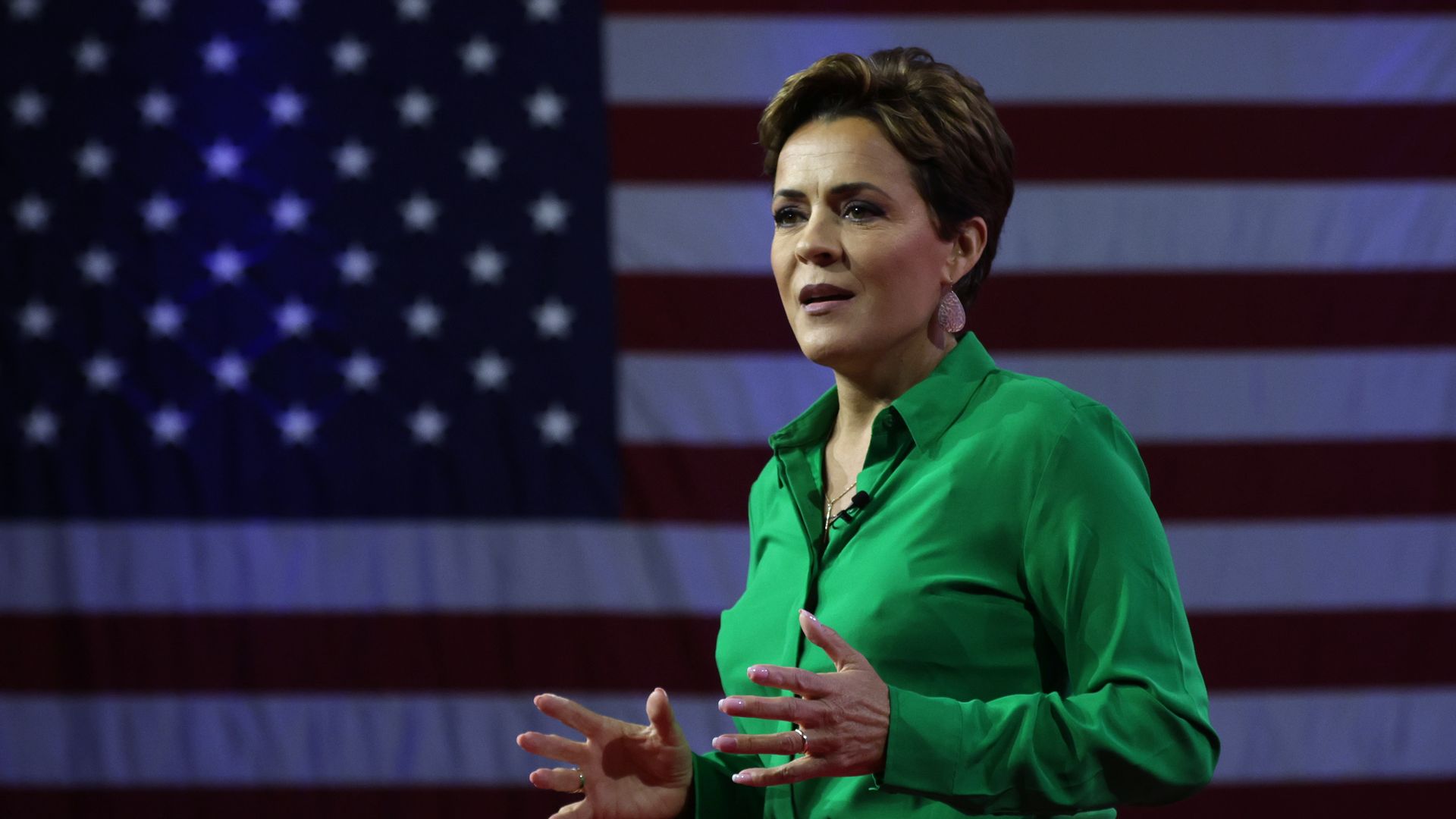 A woman gestures with her hands in front of a large United States flag.