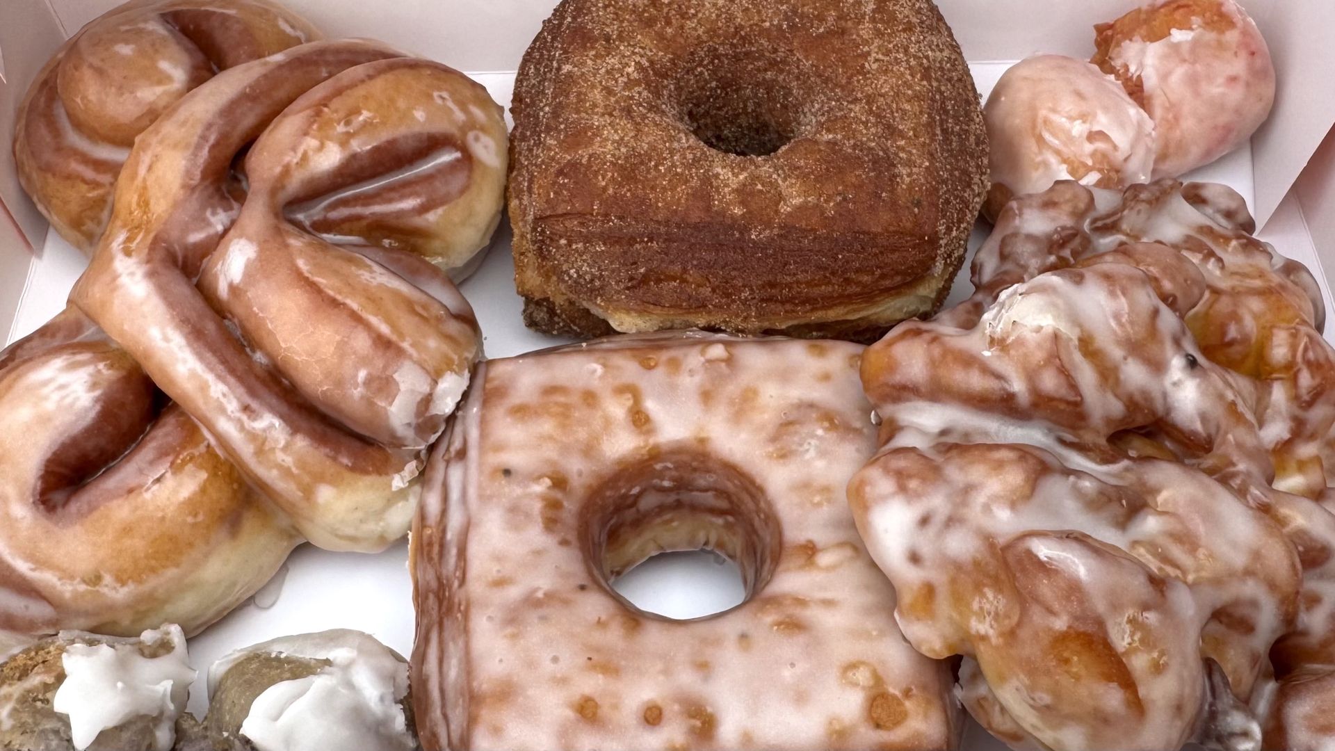 A box of assorted glazed donuts, including square, twisted, and cruller shapes, topped with icing and cinnamon sugar.