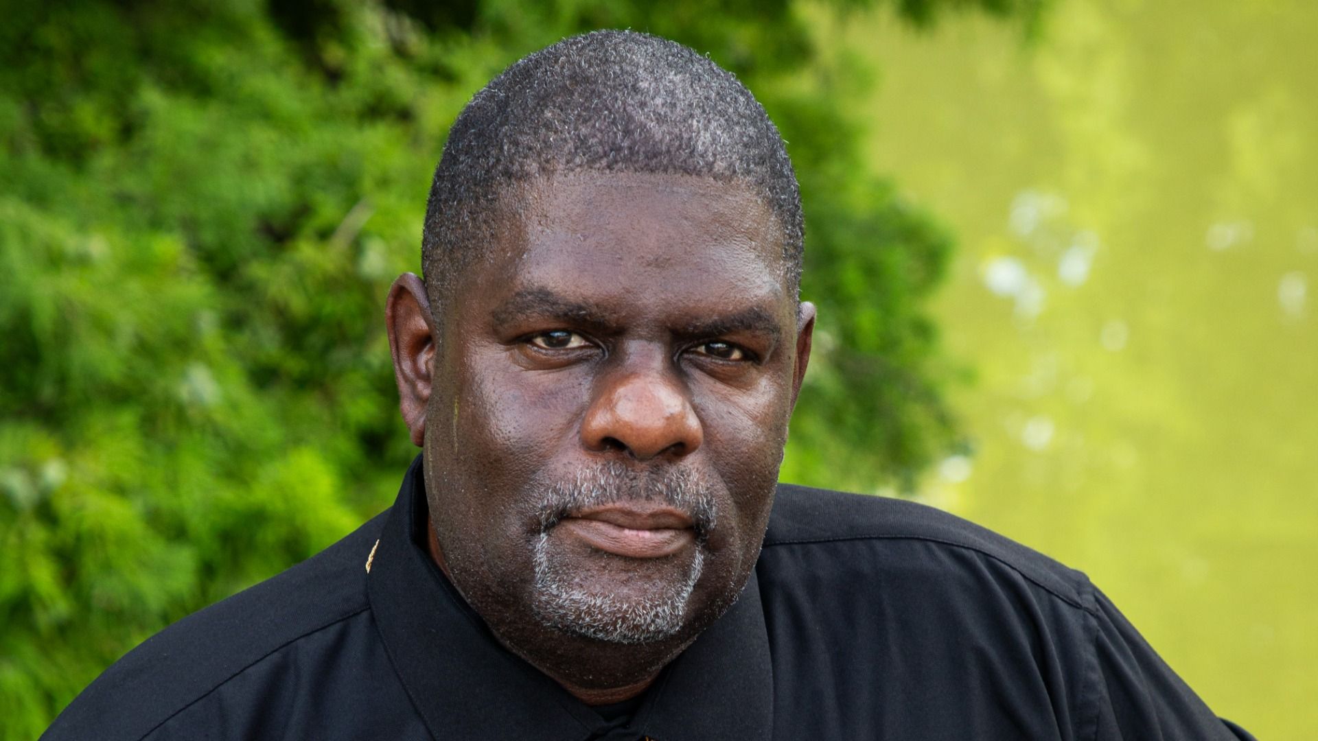 Close-up of a Black man with short gray beard, wearing a black shirt, outdoors with green foliage behind him and a yellow-green background to the right, looking at the camera.