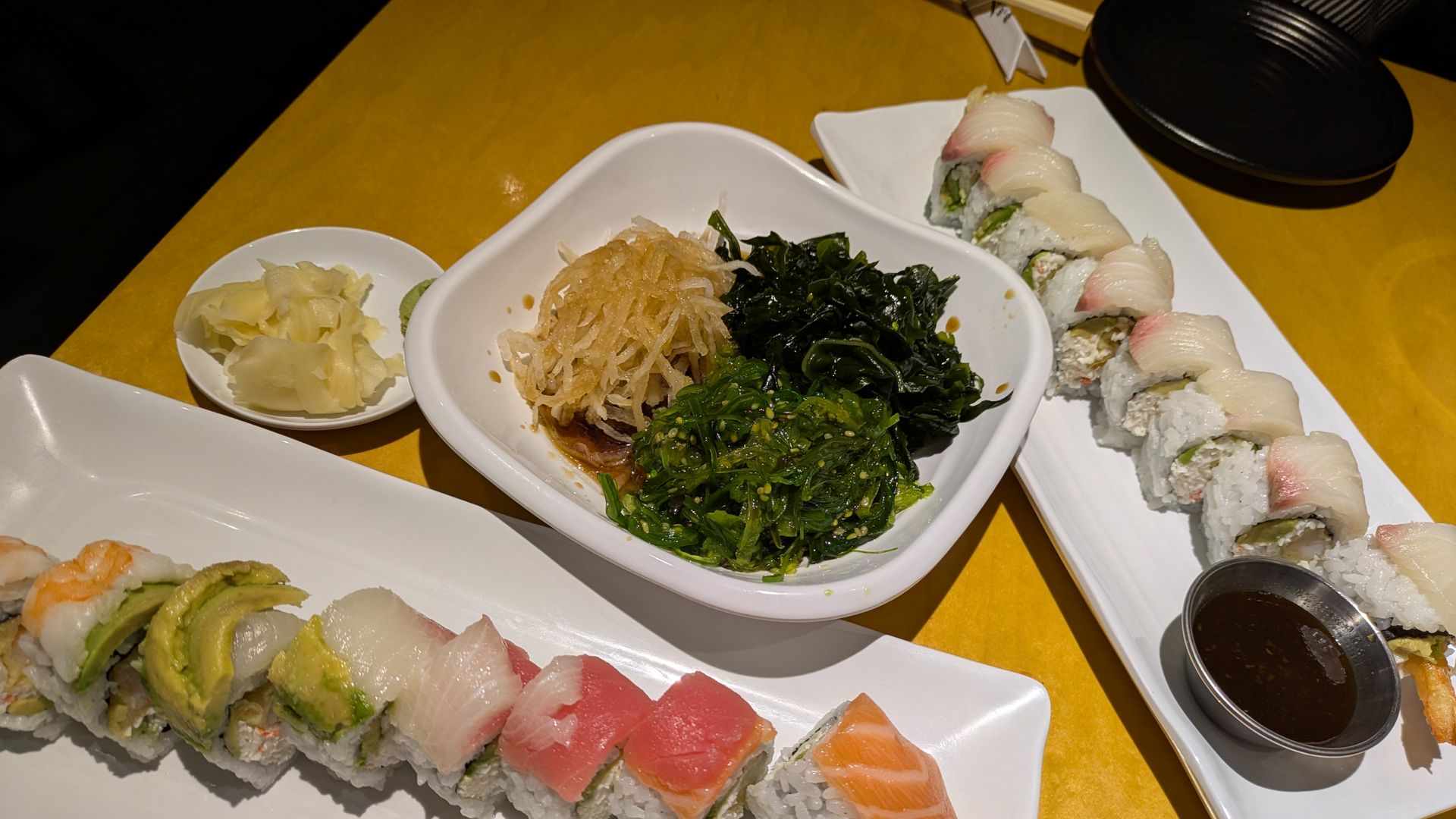 Two plates of assorted sushi rolls with avocado, fish, and tempura shrimp, a bowl of seaweed salad, pickled ginger, and a small dish of soy sauce on a yellow table.