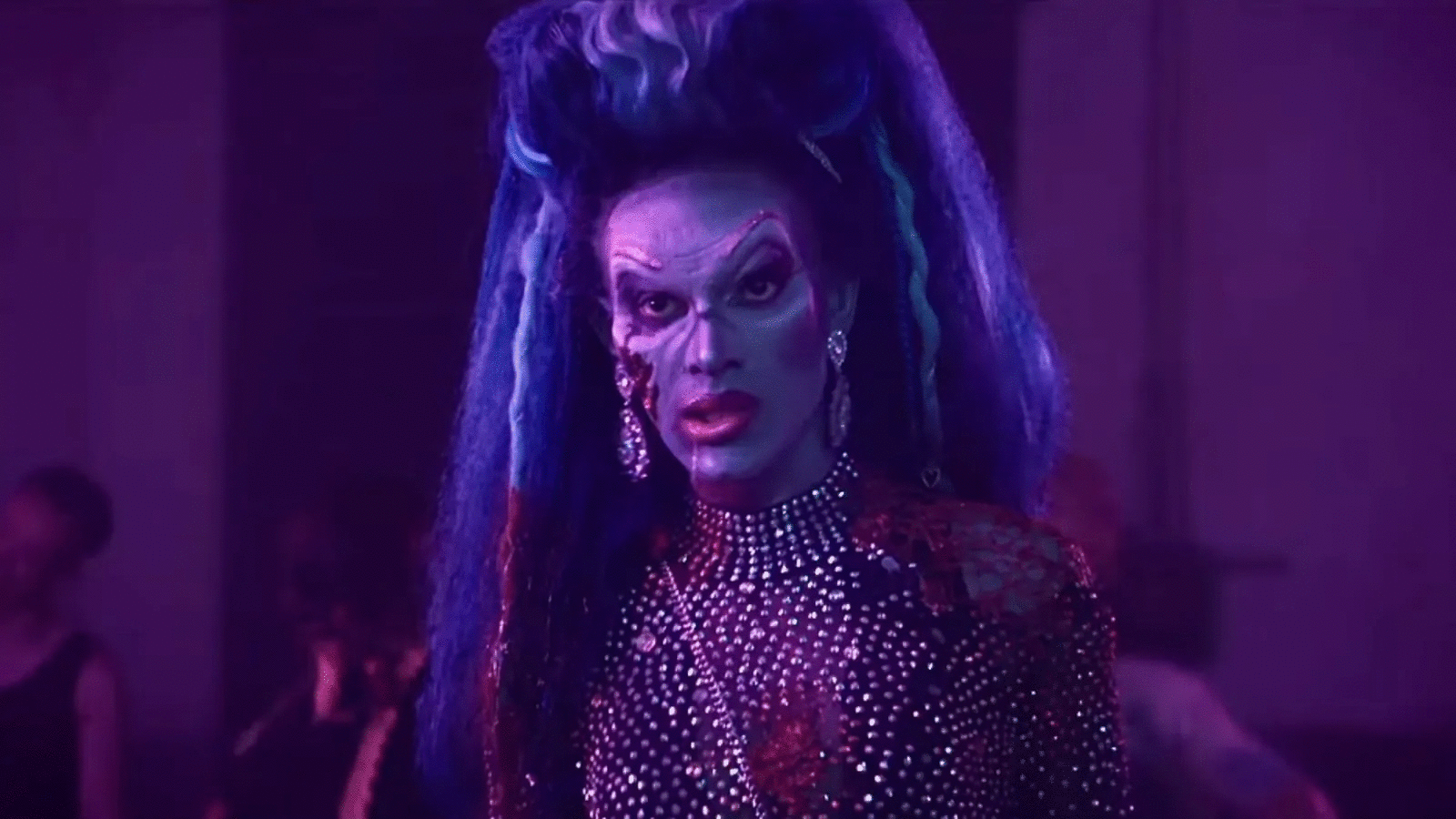 Person with dramatic purple and blue hair, white face makeup, and red lips wearing sparkling rhinestone-studded outfit and earrings, set against a purple-lit background with blurred figures.