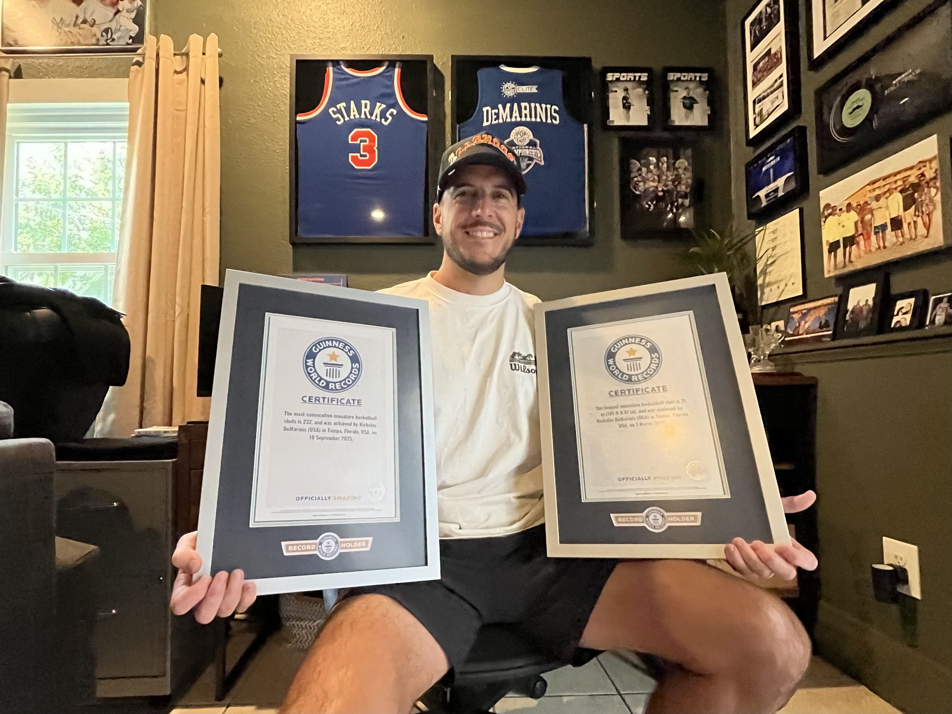 Smiling man wearing a cap sits indoors, holding three Guinness World Records certificates. Behind him, framed blue basketball jerseys read "Starks" and "DeMarinis," with photos on the wall.