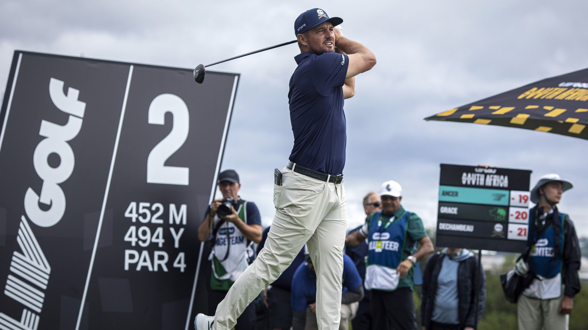 Male golfer in a navy shirt and white pants mid-swing with a driver on a golf course, near a large hole 2 sign reading 452 m, 494 y, par 4, with spectators in the background.