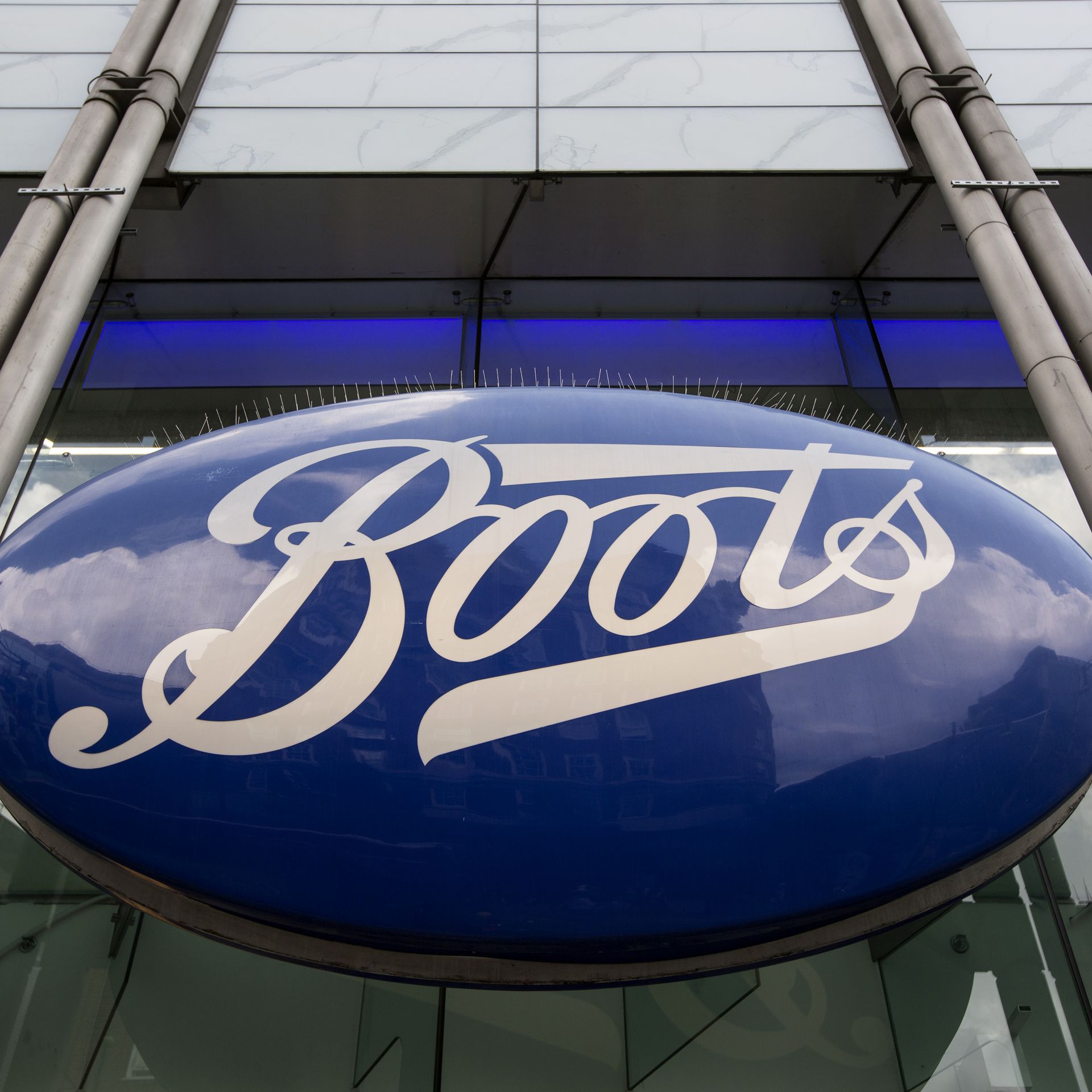 An oval sign, featuring the pharmacy chain Boots' name spelled out in white lettering on a blue background, hangs off the side of a glass-covered building.
