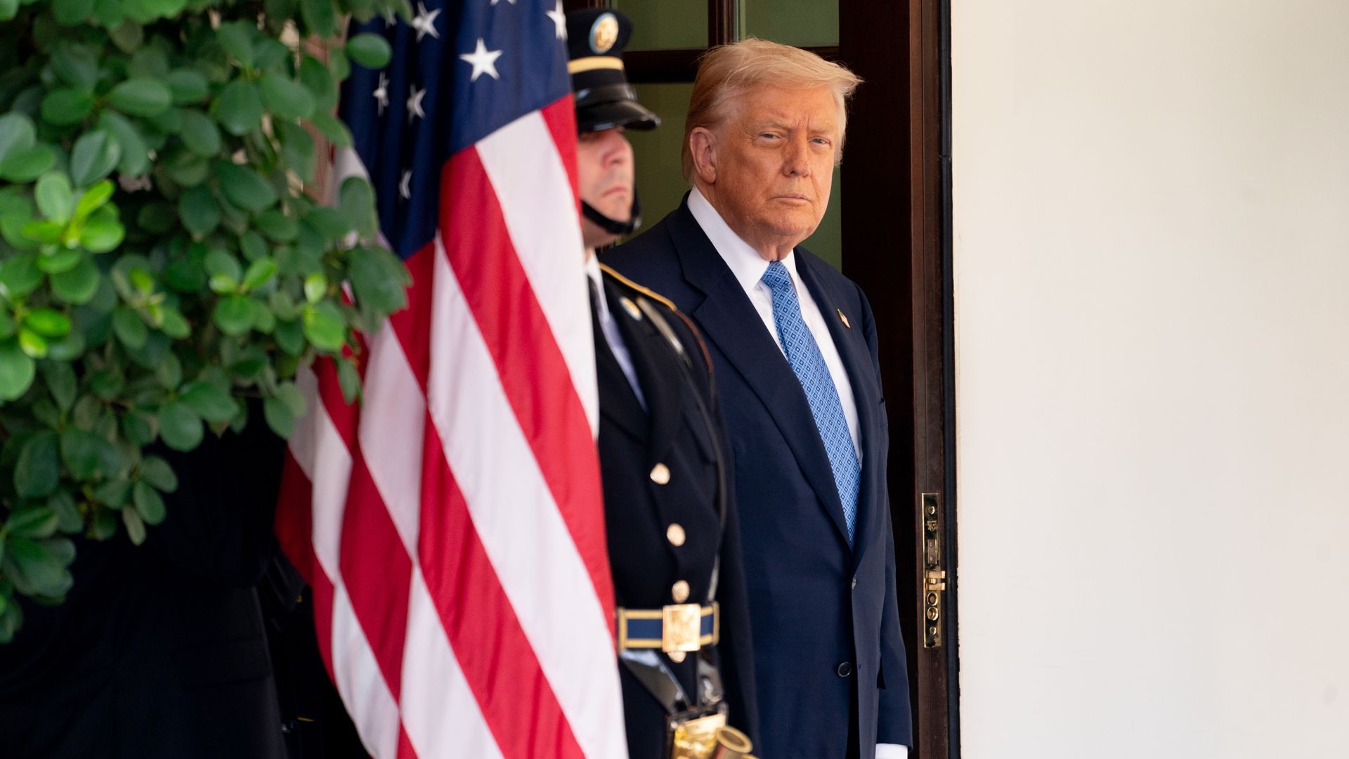 A man in a dark suit and blue patterned tie stands at a doorway next to a uniformed guard holding a U.S. flag, with green leaves partially visible on the left.