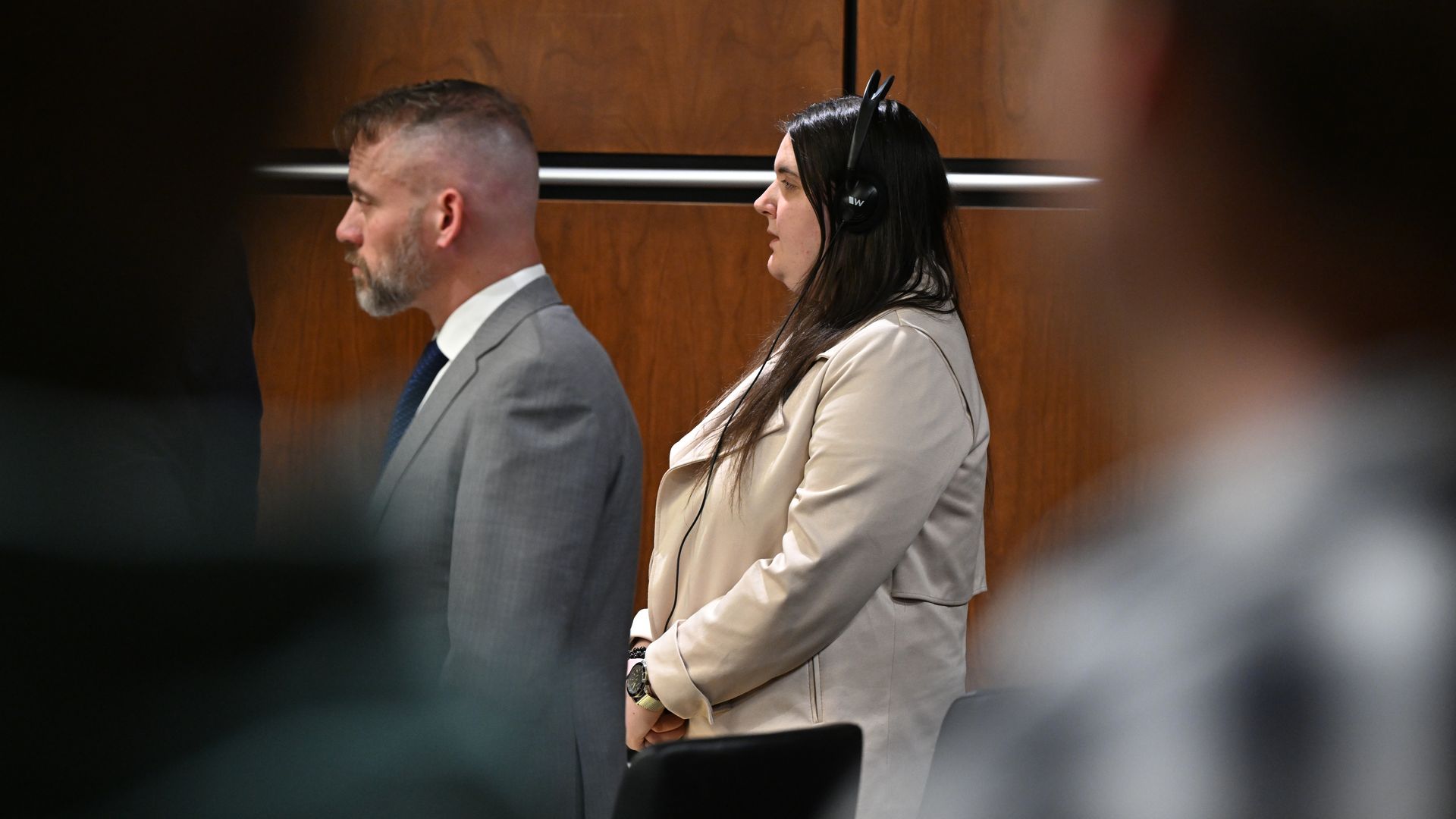 Side view of Yeva Smilianska wearing a beige jacket and headphones standing next to a man with short hair, beard, and a gray suit in a wood-paneled room.