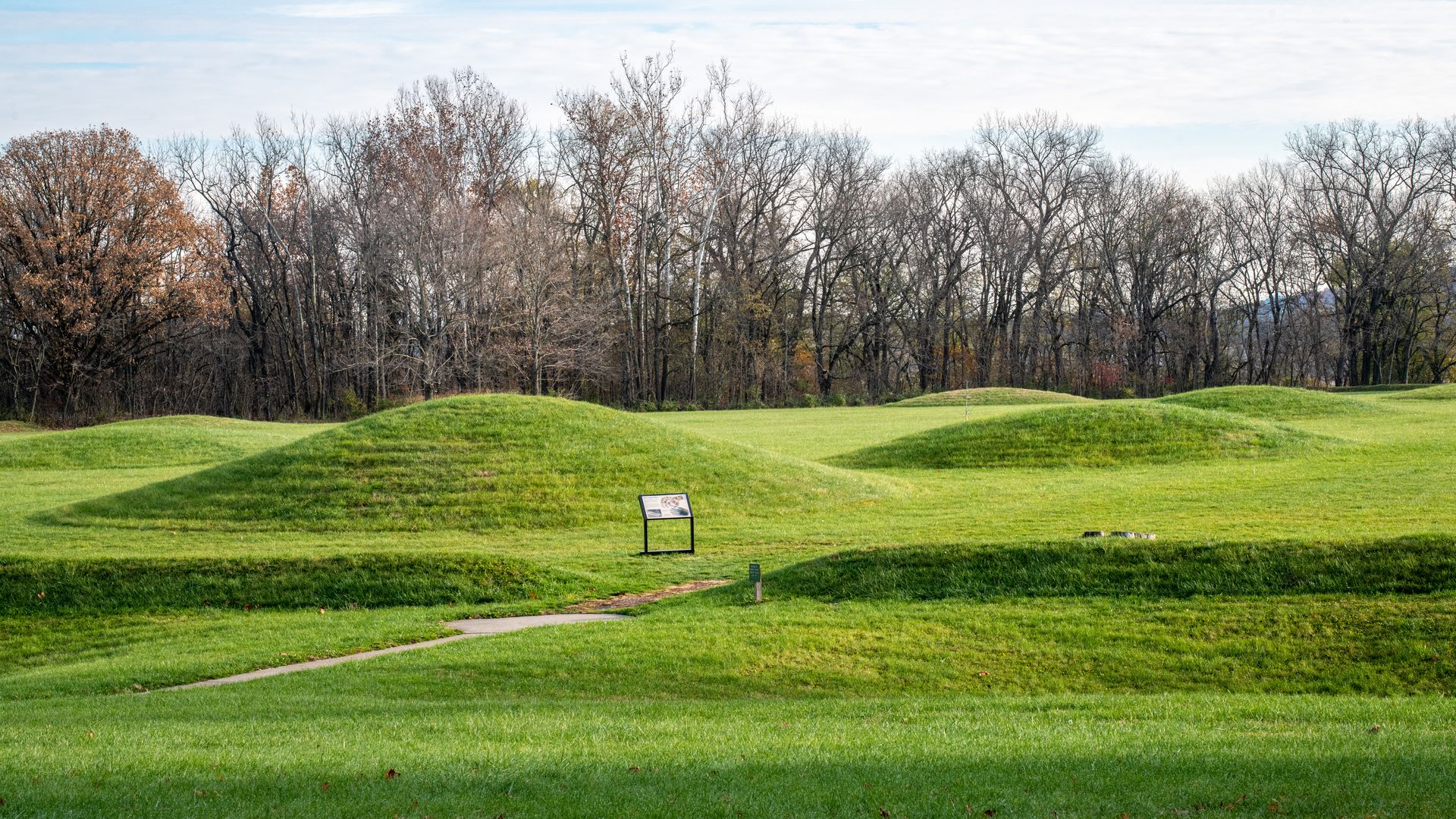 Green grassy mounds in a park with a path and an informational sign, surrounded by leafless trees under a partly cloudy sky.