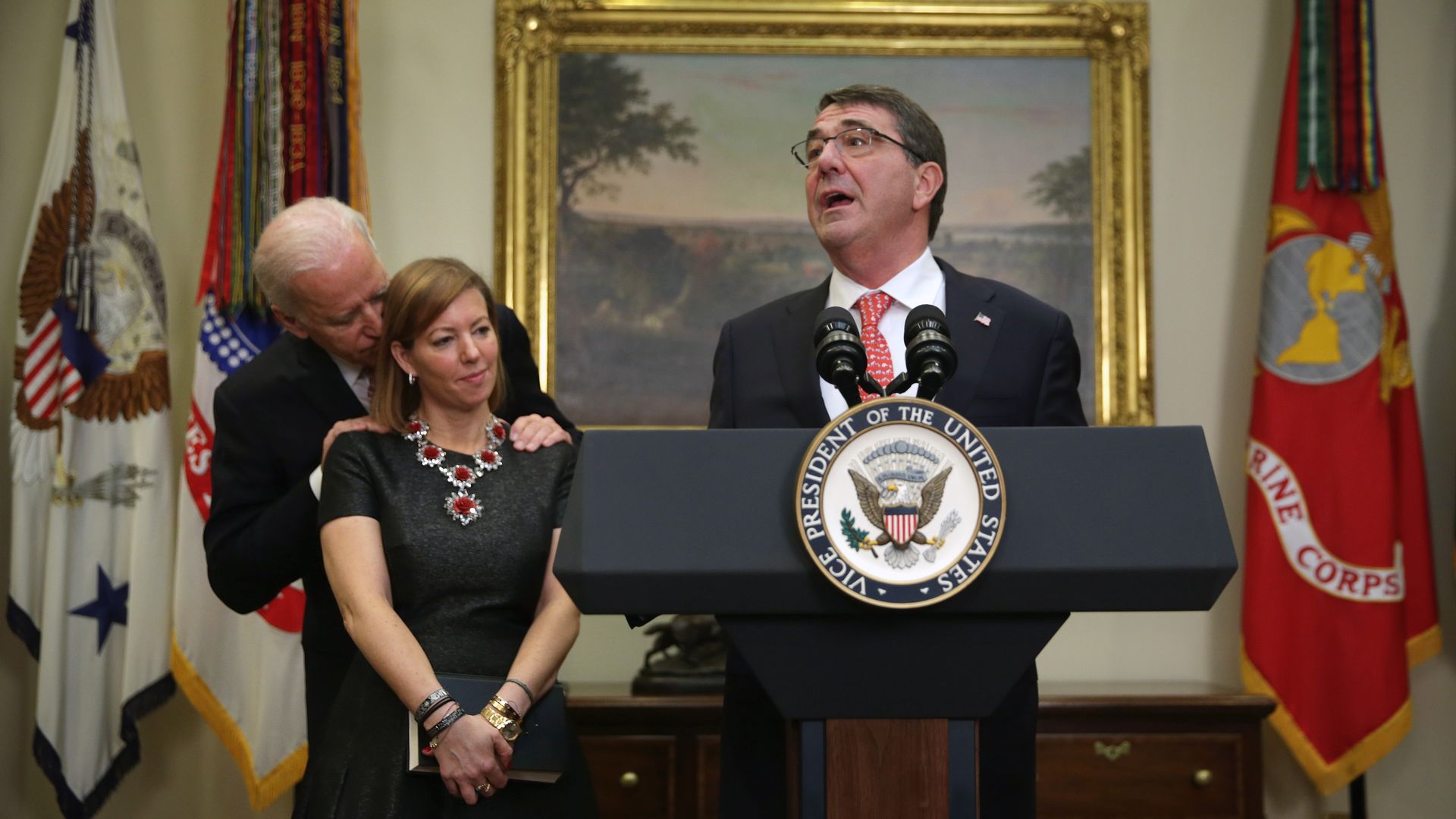 Joe Biden with hands on Stephanie Carter's shoulders, while Ash Carter speaks at podium.