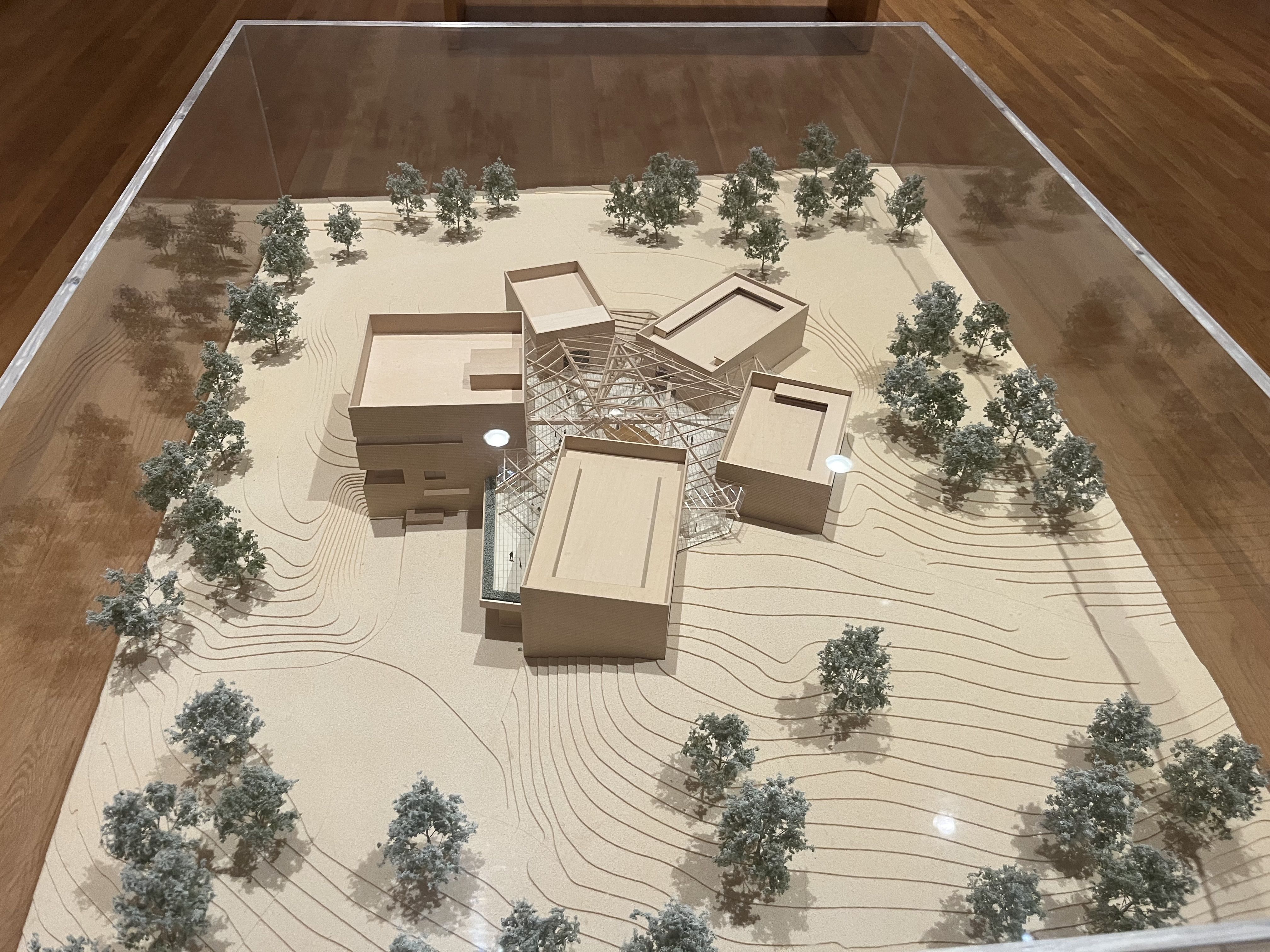 Architectural scale model in a glass display case, beige buildings around a central glass-and-steel atrium, contour lines on the base, and numerous small trees surrounding the site.