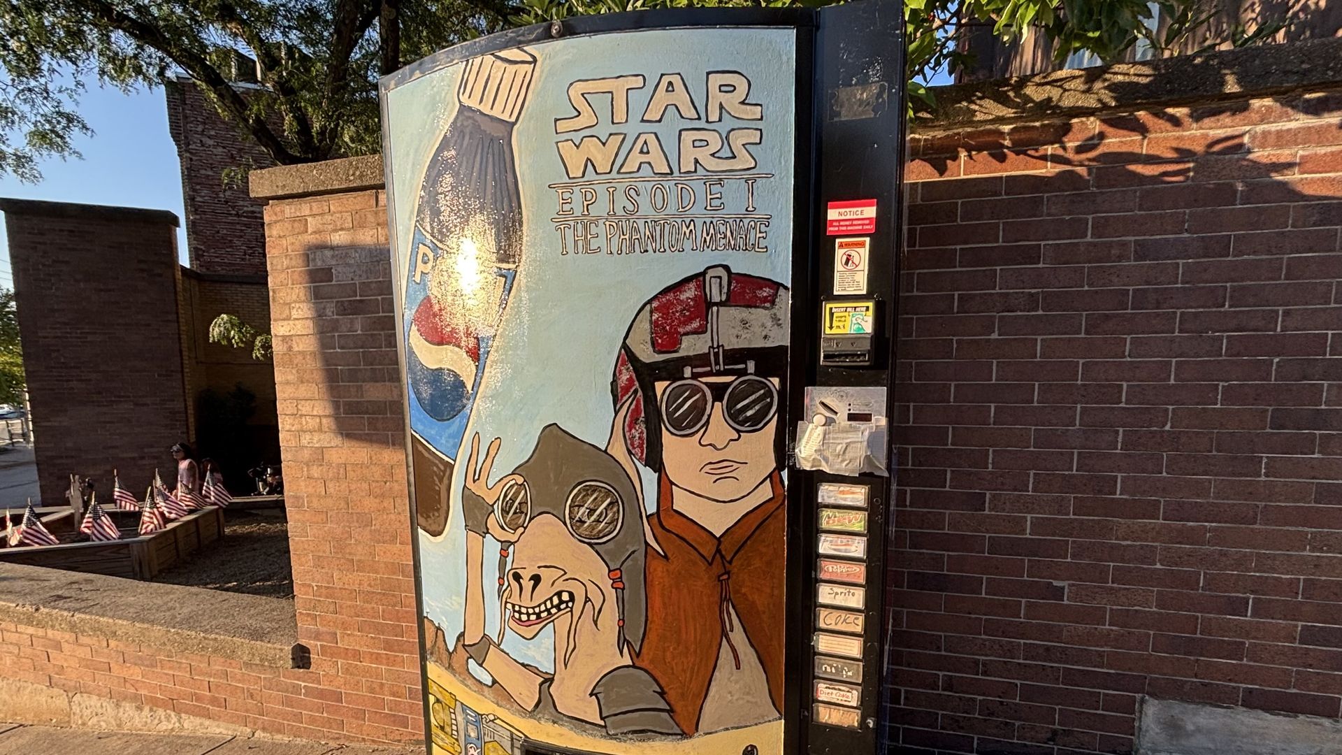 Vending-kiosk on a brick wall displays a Star Wars Episode I: The Phantom Menace poster with stylized characters and sunglasses on a blue background; small American flags line a nearby railing.