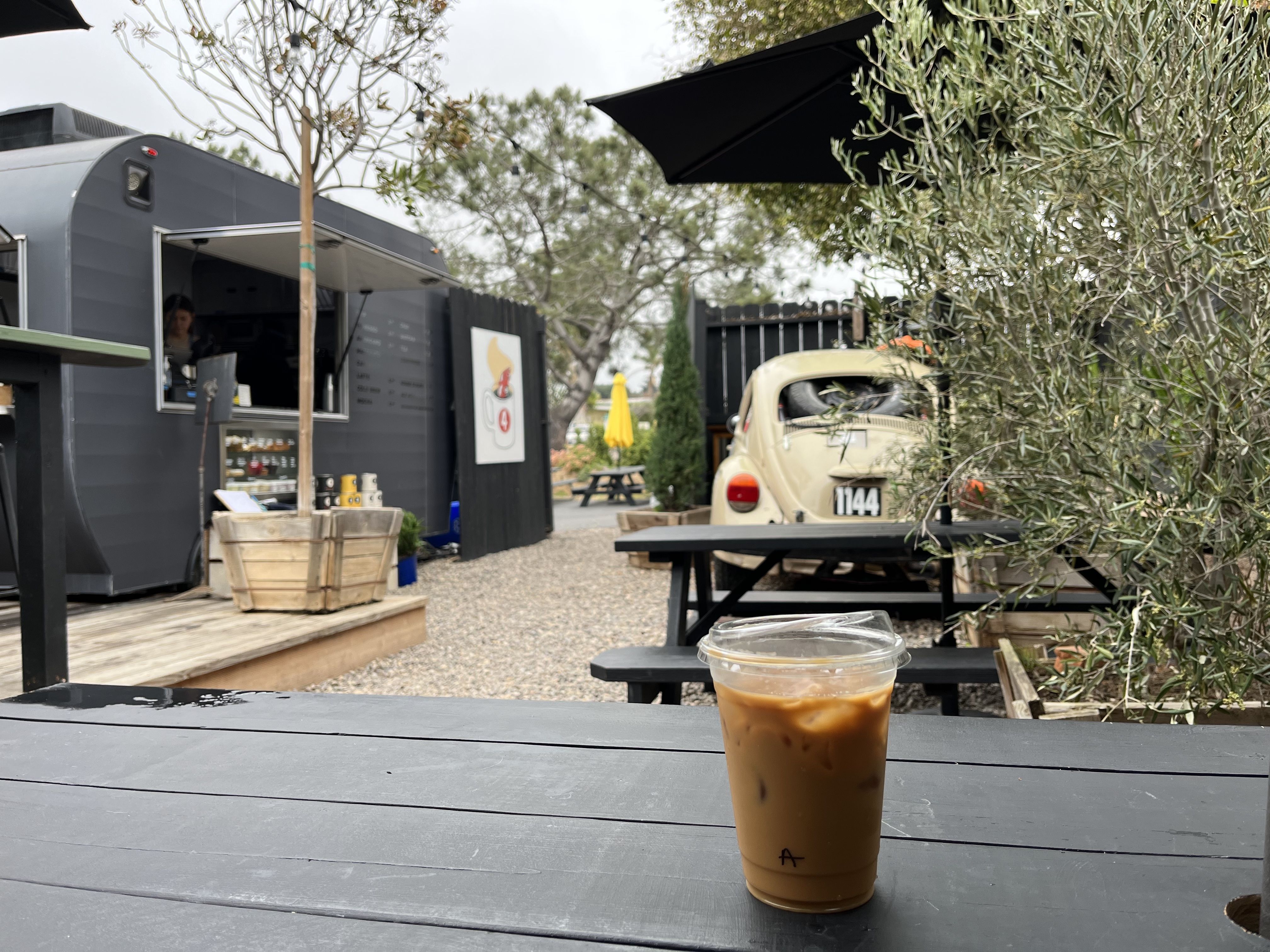 An iced coffee sits on a black picnic table at an outdoor coffee shop that features a beige classic car.  