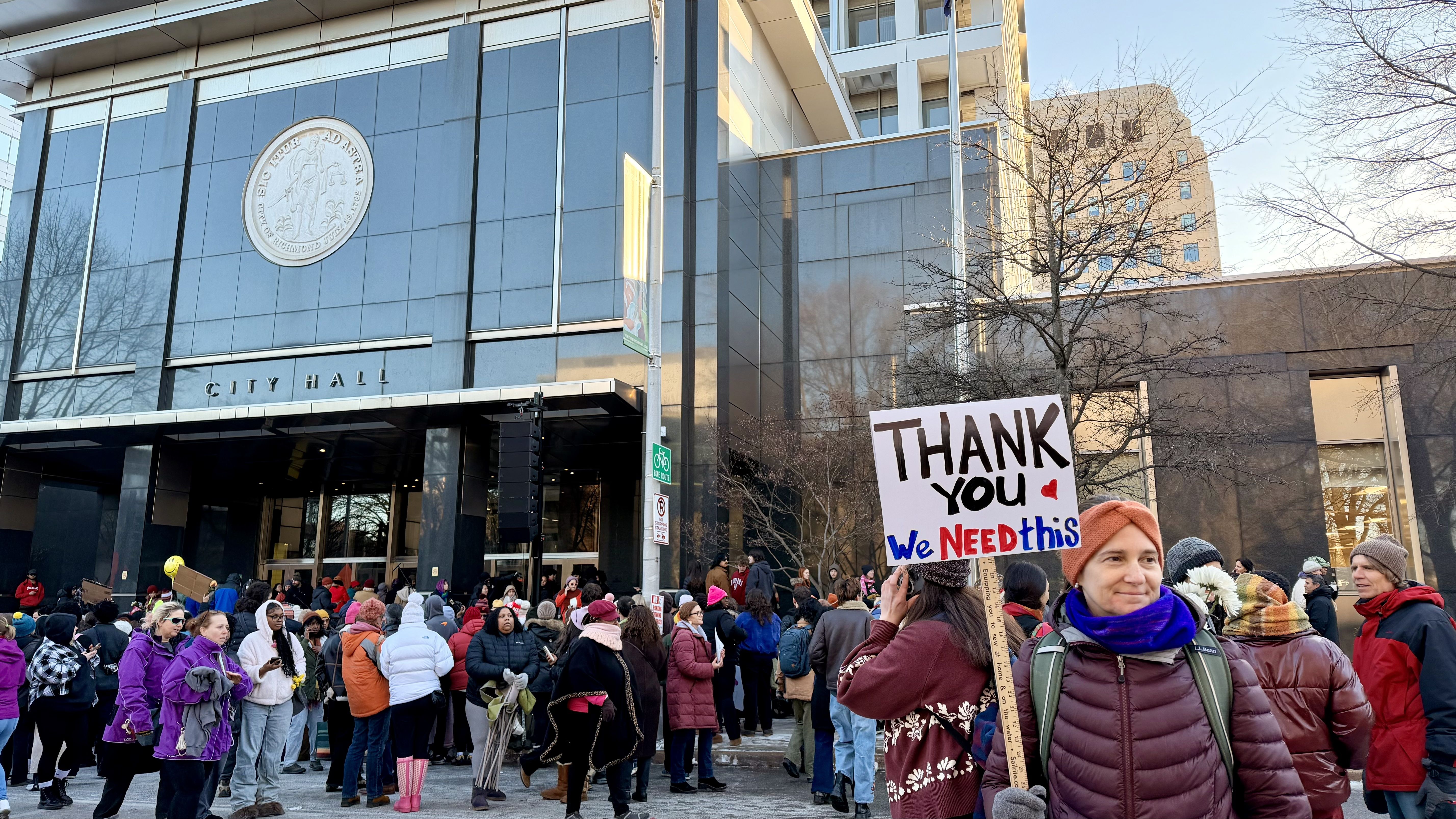 Crowd of people gathered outside a glass City Hall building on a cold day, some wearing colorful winter jackets. One person holds a sign reading, "THANK YOU We NEED this."
