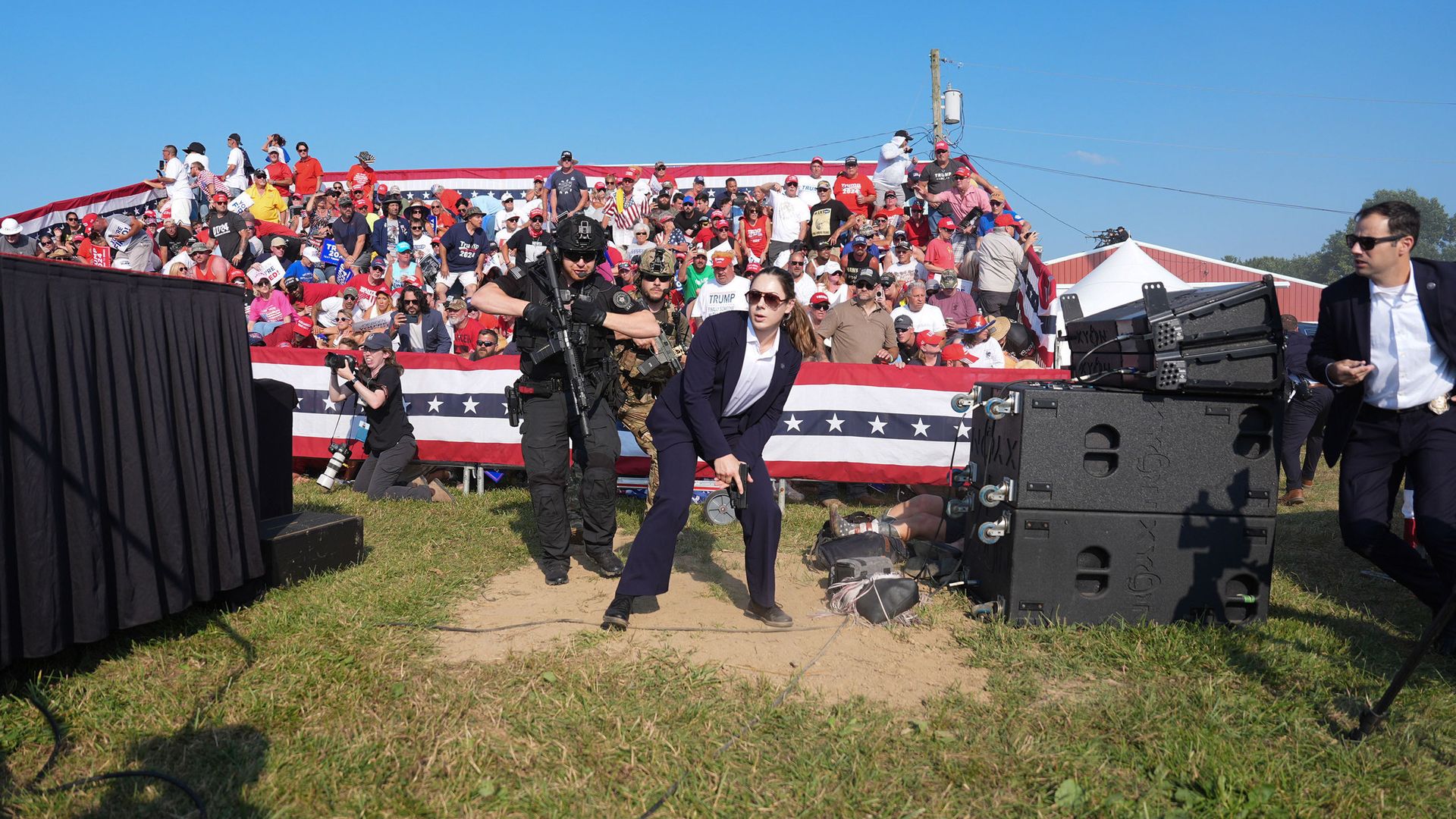 A woman wearing a suit and carrying a gun crouches on a patch of dirt, with people carrying machine guns behind her. A crowd of people on bleachers are in the background.