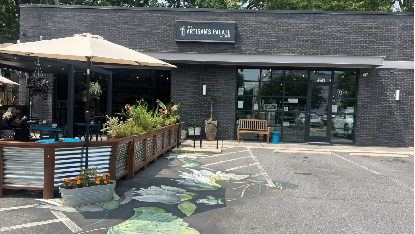 Brick storefront reading "The Artisan's Plate" with outdoor seating under a beige umbrella, wooden railing, potted flowers, and a large leaf mural on the pavement; glass entry doors on the right.