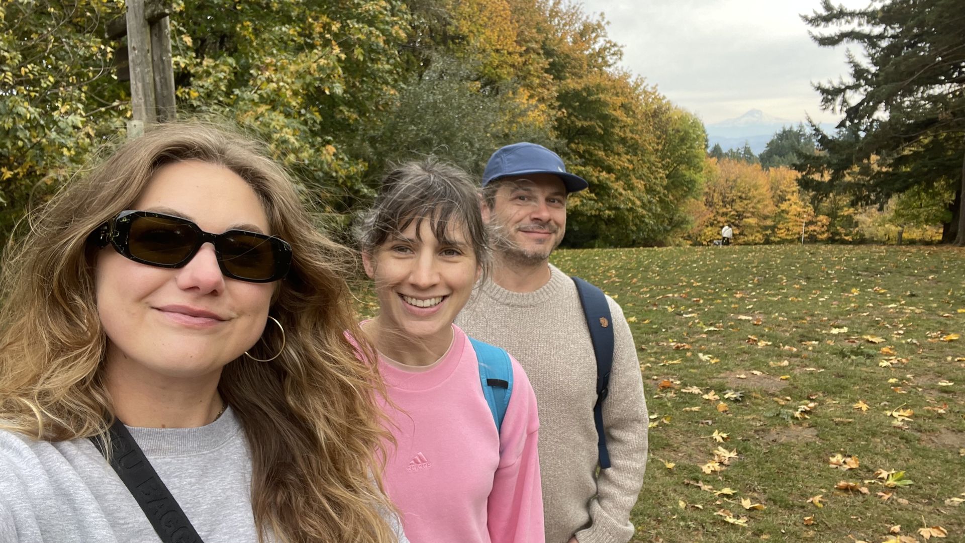 Three people smiling outdoors in autumn; woman on left with sunglasses and curly hair, woman in middle wearing pink Adidas sweater, man on right with blue cap and beige sweater, colorful fall trees behind.