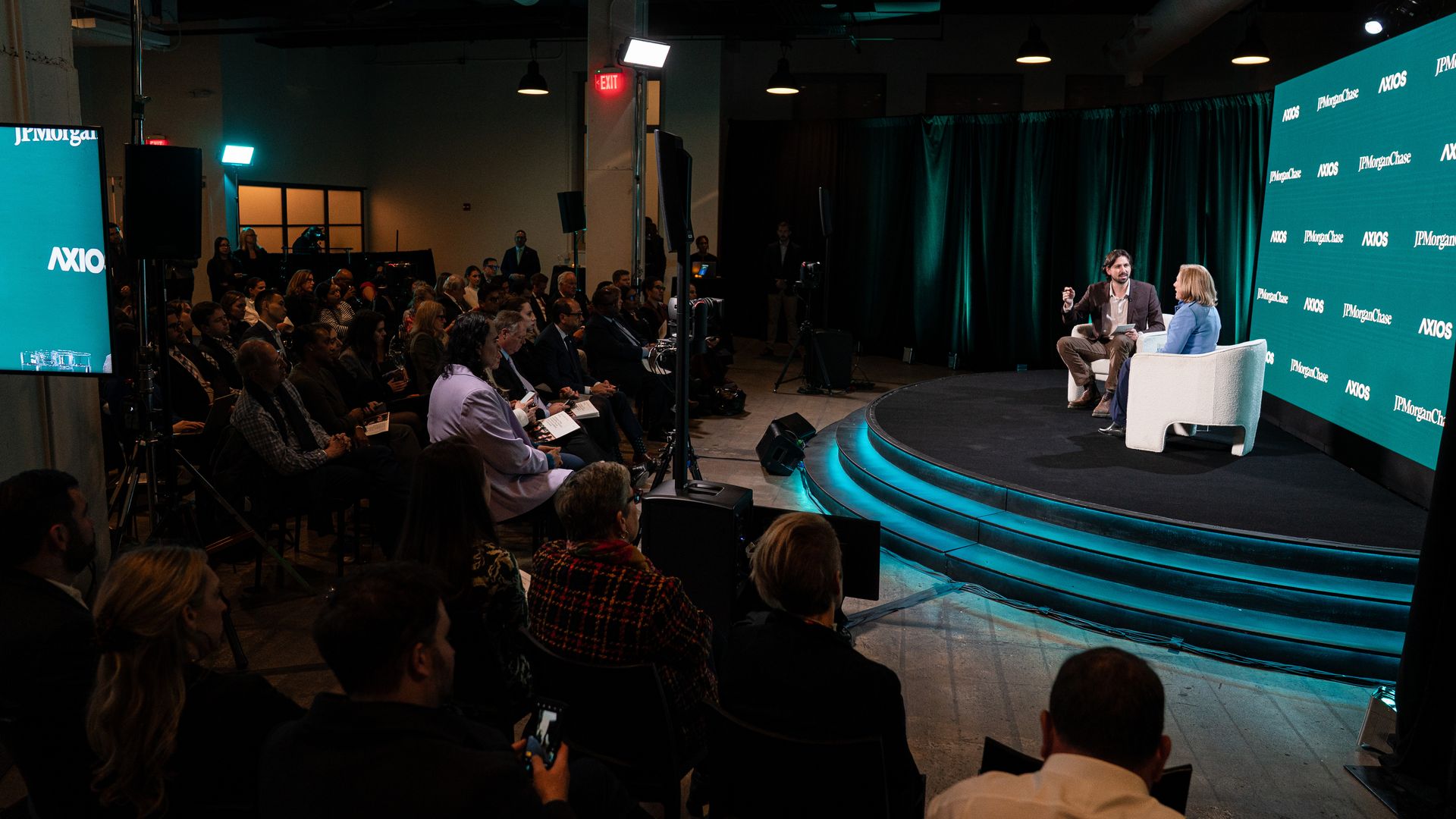 A panel discussion with two speakers seated on white chairs on a stage with curved steps. The background shows logos for JPMorgan Chase and Axios. Audience listens attentively.