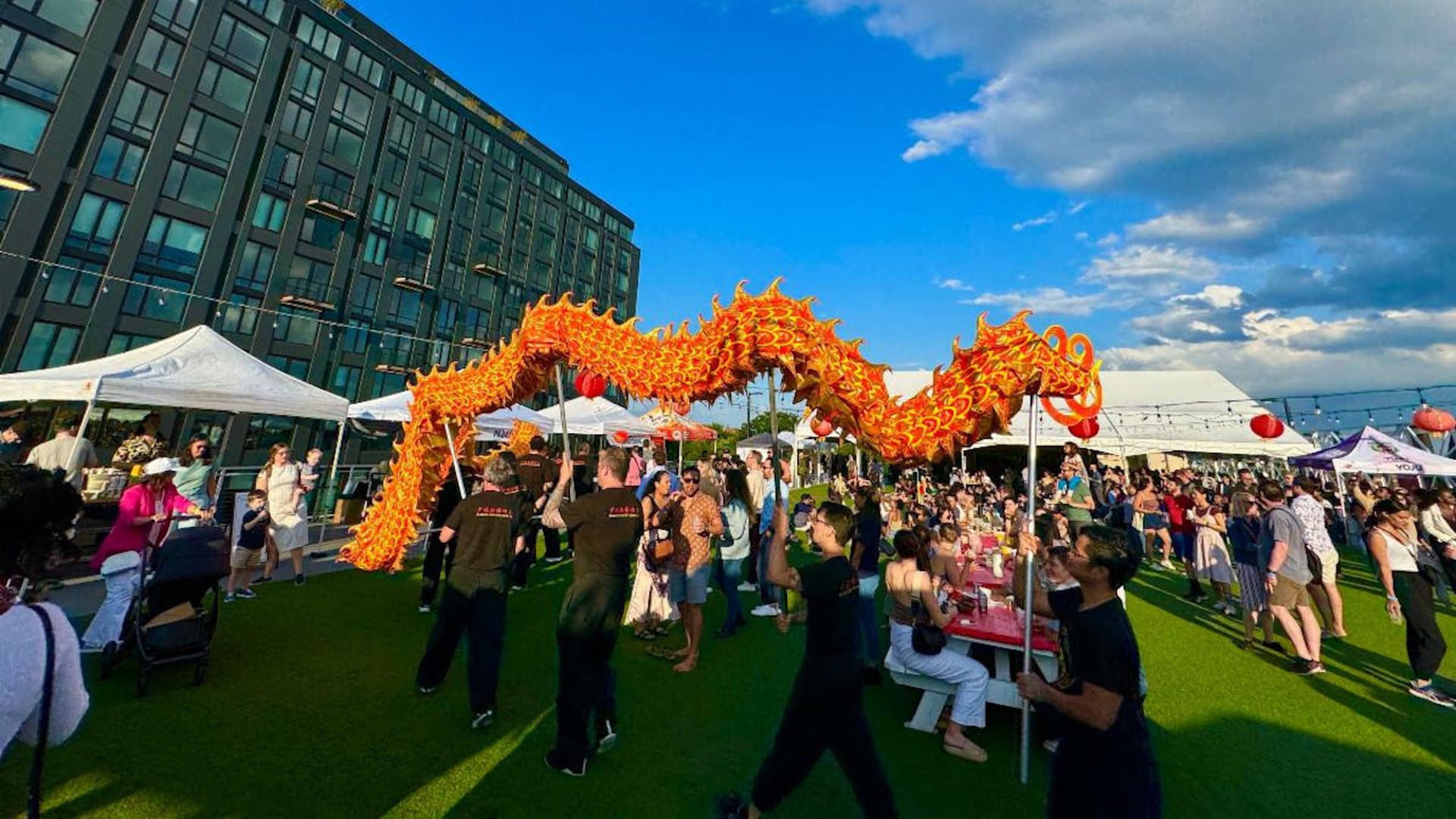 An outdoor festival on a grassy plaza features a bright orange dragon dance weaving over a crowd beneath white tents; a modern glass building sits to the left, with a blue sky overhead.