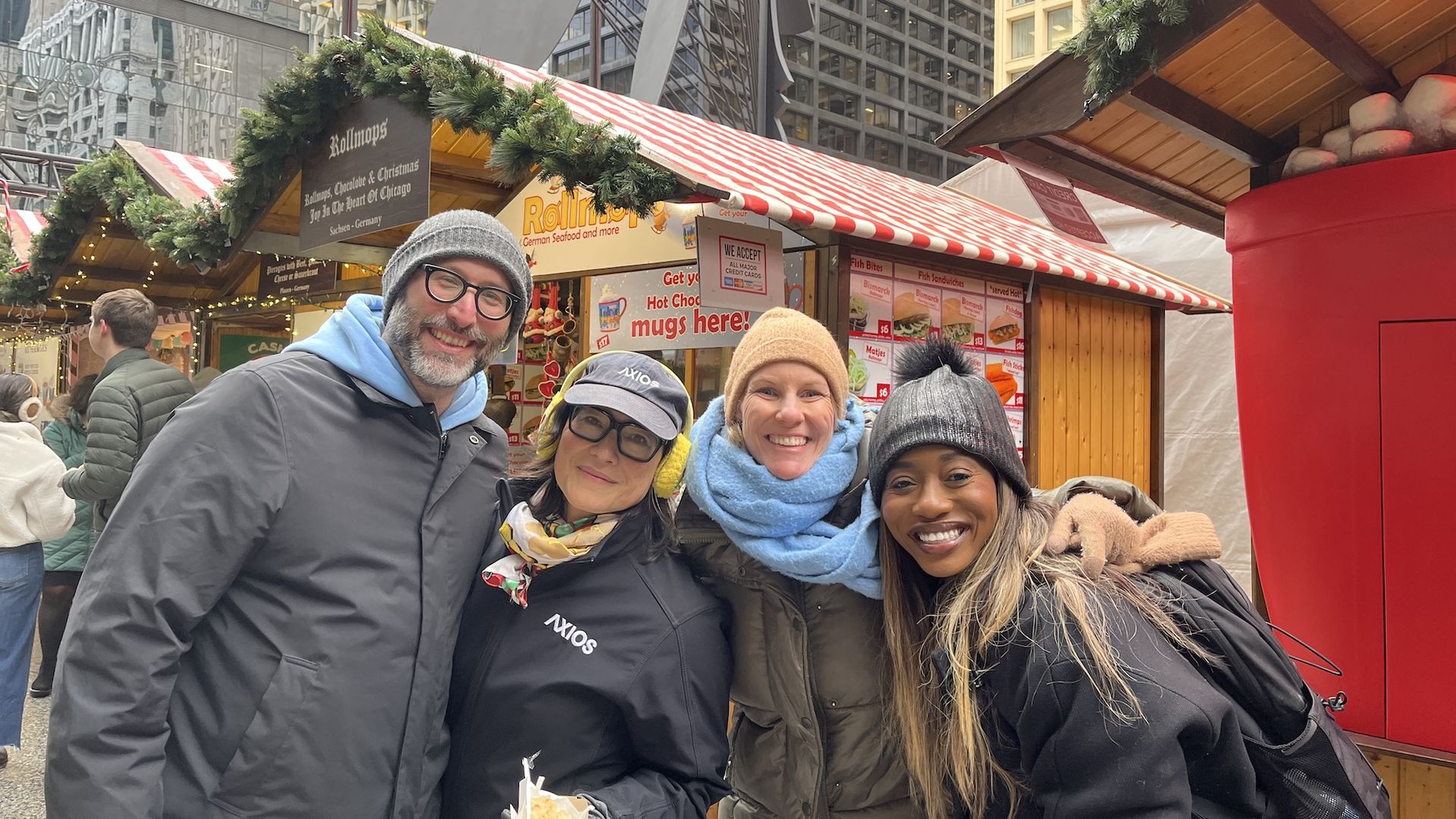 Justin Kaufmann, Monica Eng, Carrie Shepherd, Moyo Adeolu  smiling at Chicago's Christkindlmarket holiday market with decorated stalls and menus, dressed in winter coats, hats, and scarves, standing close together outdoors in a city.