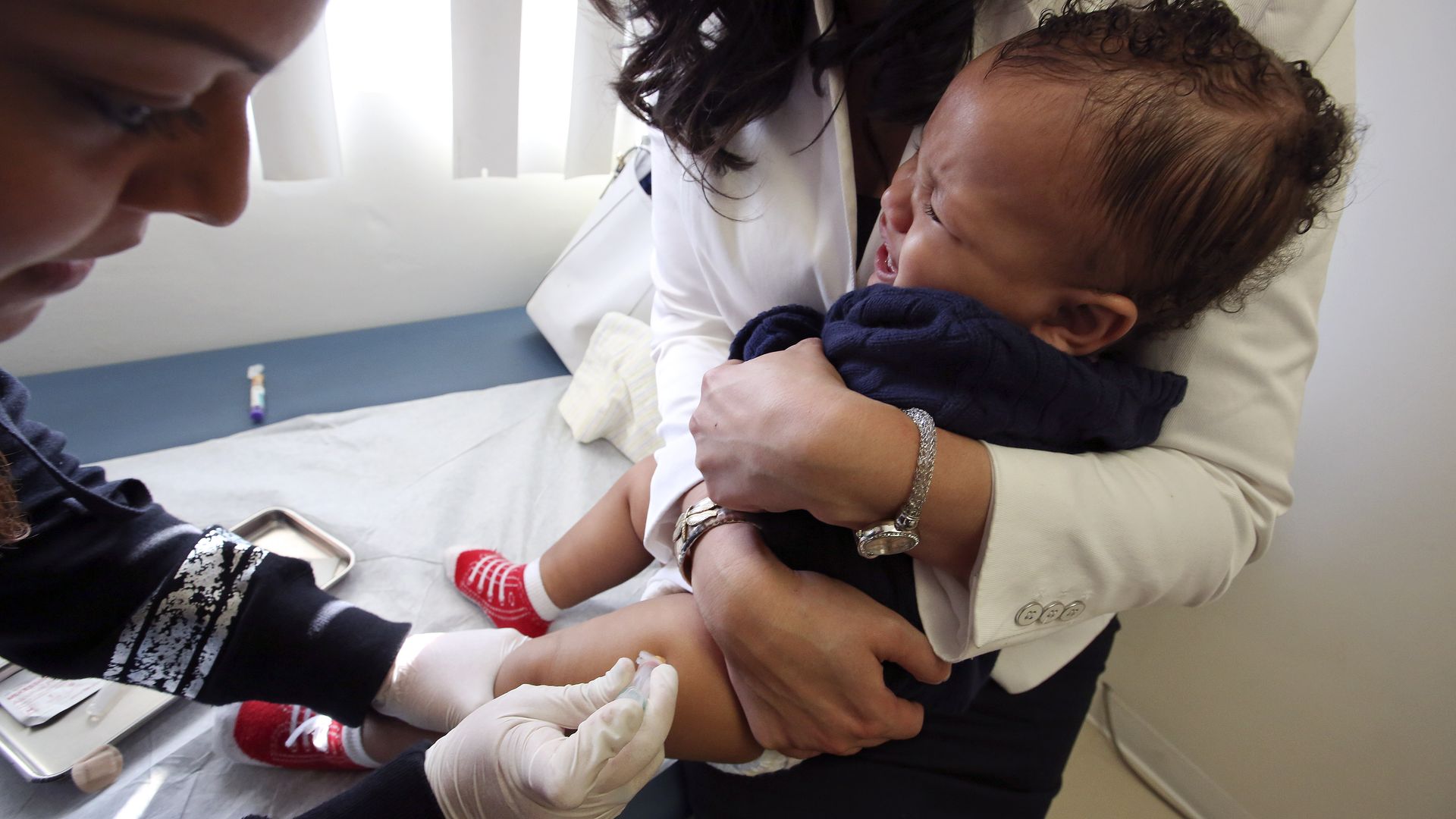 In this image, a woman holds a baby as it is given a shot by a medical professional.