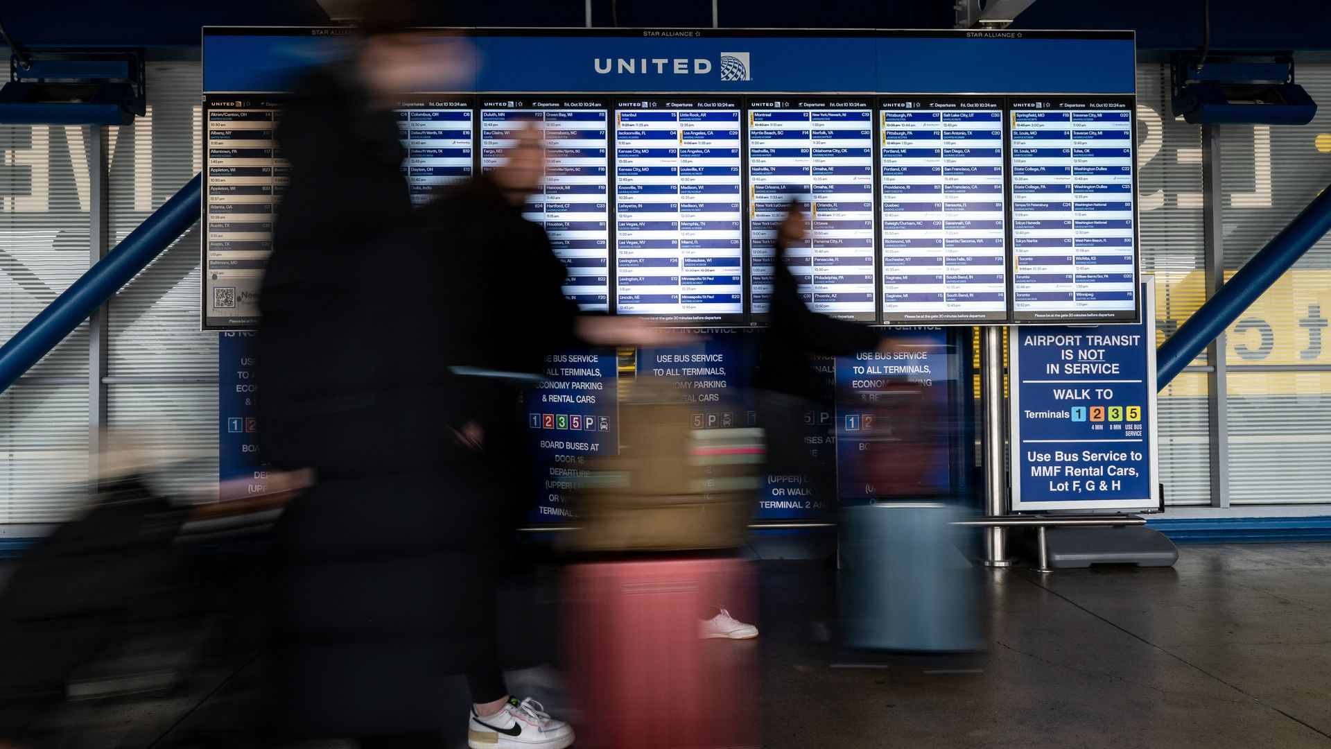 A blurry photo of passengers at an airport walking past a board of flight information.