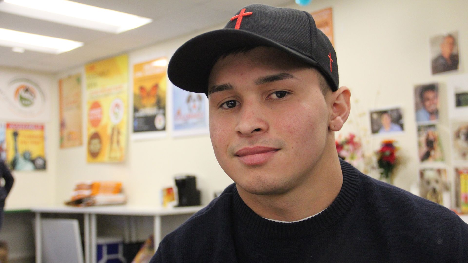 A man in a white sweater and a black hat with a red cross looks on, while a white wall with numerous and colorful posters are placed.