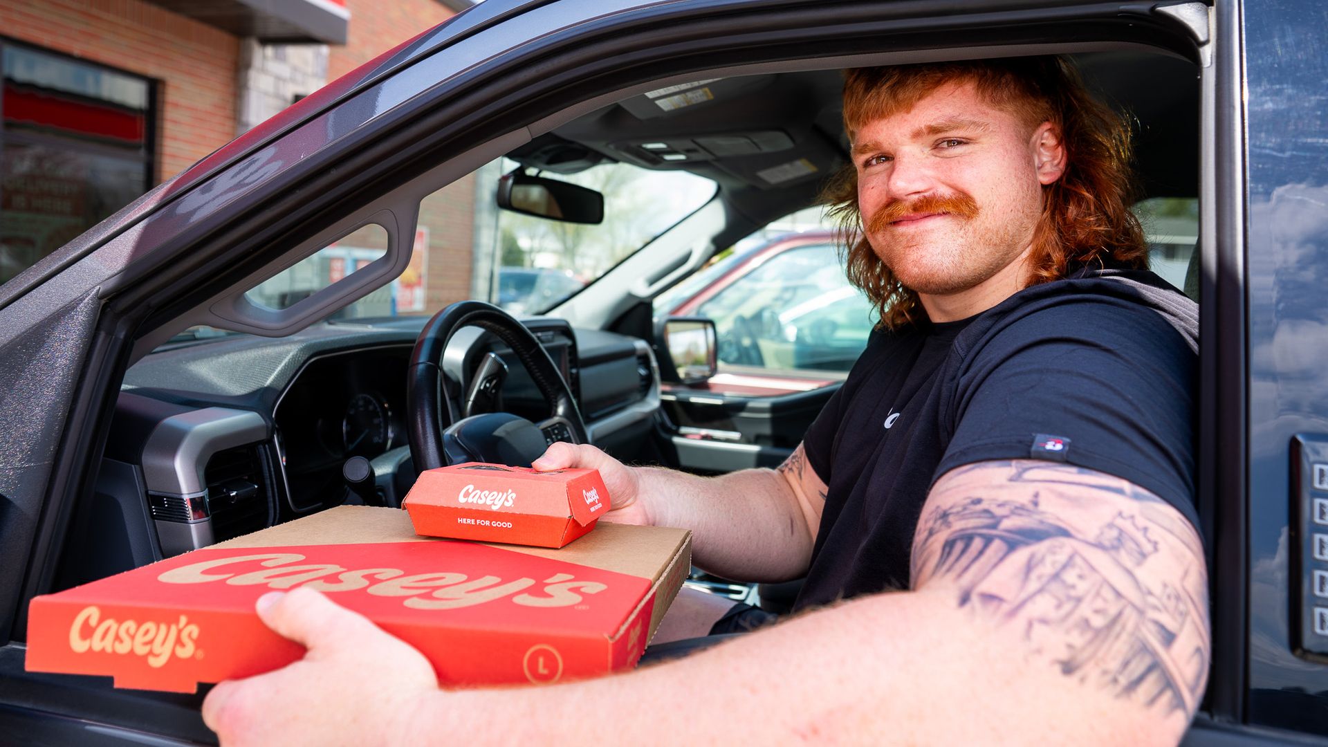 Gennings Dunker with red hair and a mustache sits in a pickup window, handing a Casey's orange pizza box to the camera; other Casey's boxes are visible in the foreground.