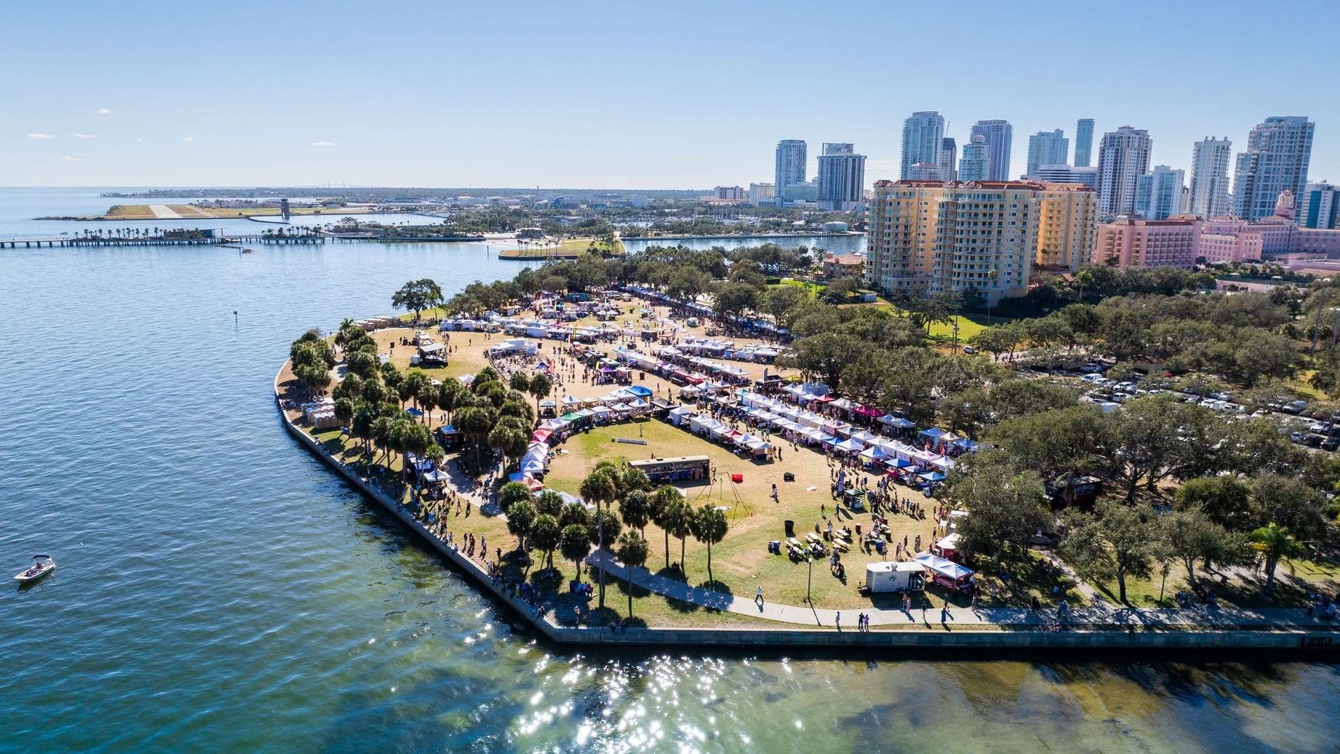 Aerial view of a sunny coastal city: blue bay on the left, a long pier, a park with many white tents and people, and a skyline of tall glass towers along the waterfront under a clear blue sky.