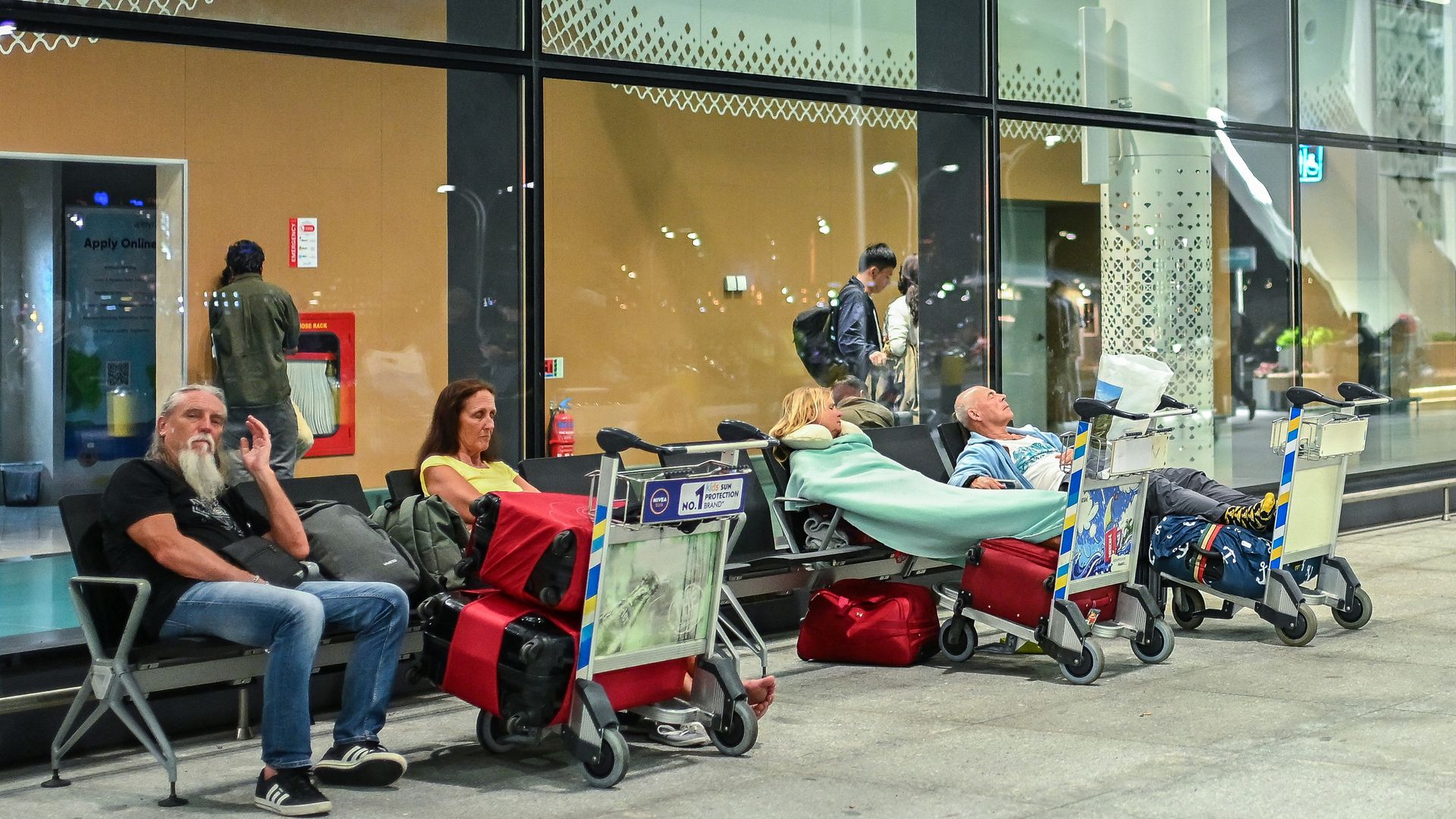 People waiting at night outside a building with glass windows, sitting on benches with luggage carts; one woman is wrapped in a green blanket, others appear tired or resting.