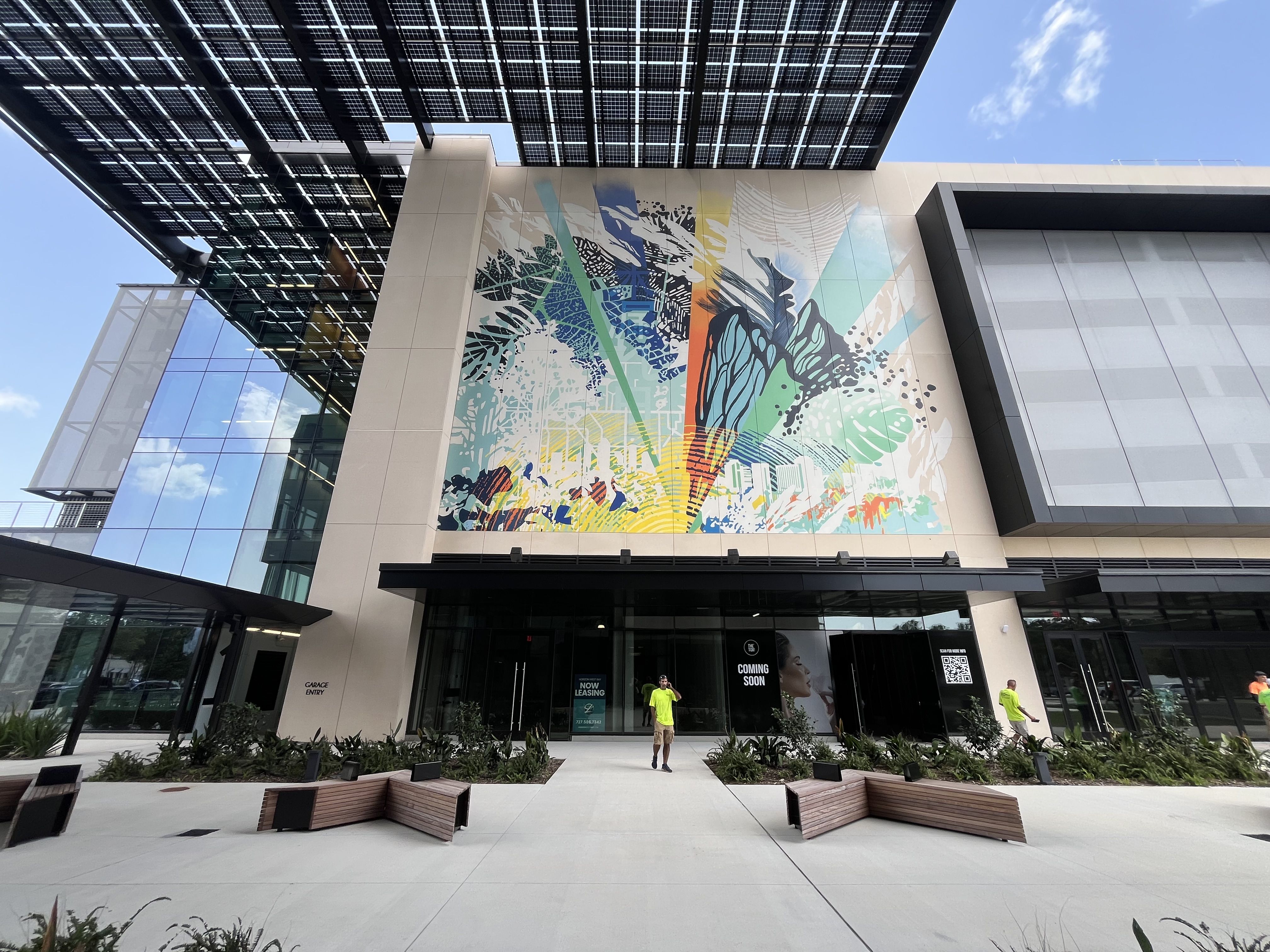 Modern building entrance with a large colorful abstract mural on a beige wall, glass windows, and a black grid canopy with solar panels overhead. Two people in neon shirts stand near the entrance; blue sky.