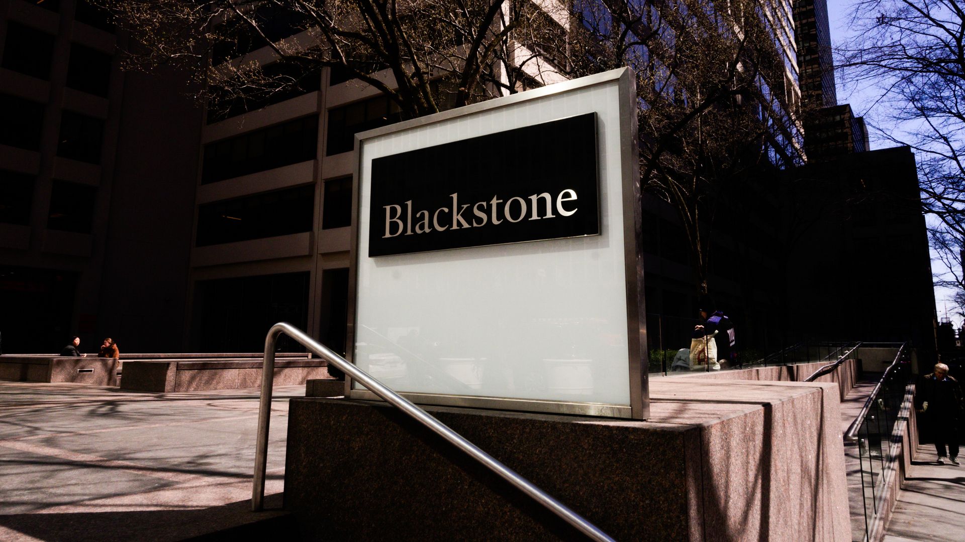 Urban plaza with a large Blackstone sign in a glass panel, tall modern buildings, leafless trees, and a blue sky; a few people sit on benches in the foreground.